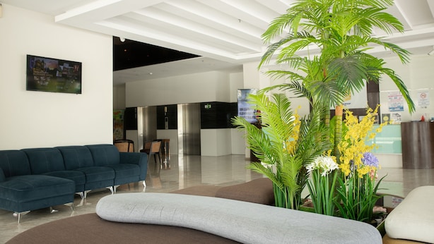 A modern lobby with a sectional sofa, a grey pouf, and a large potted plant with yellow flowers. - Golden Roof Hotel Ampang Ipoh (Ipoh)