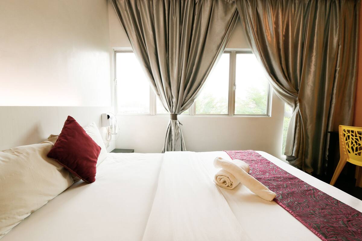 A hotel room with a bed, pillows, a rolled towel, a patterned blanket, and a window with curtains.