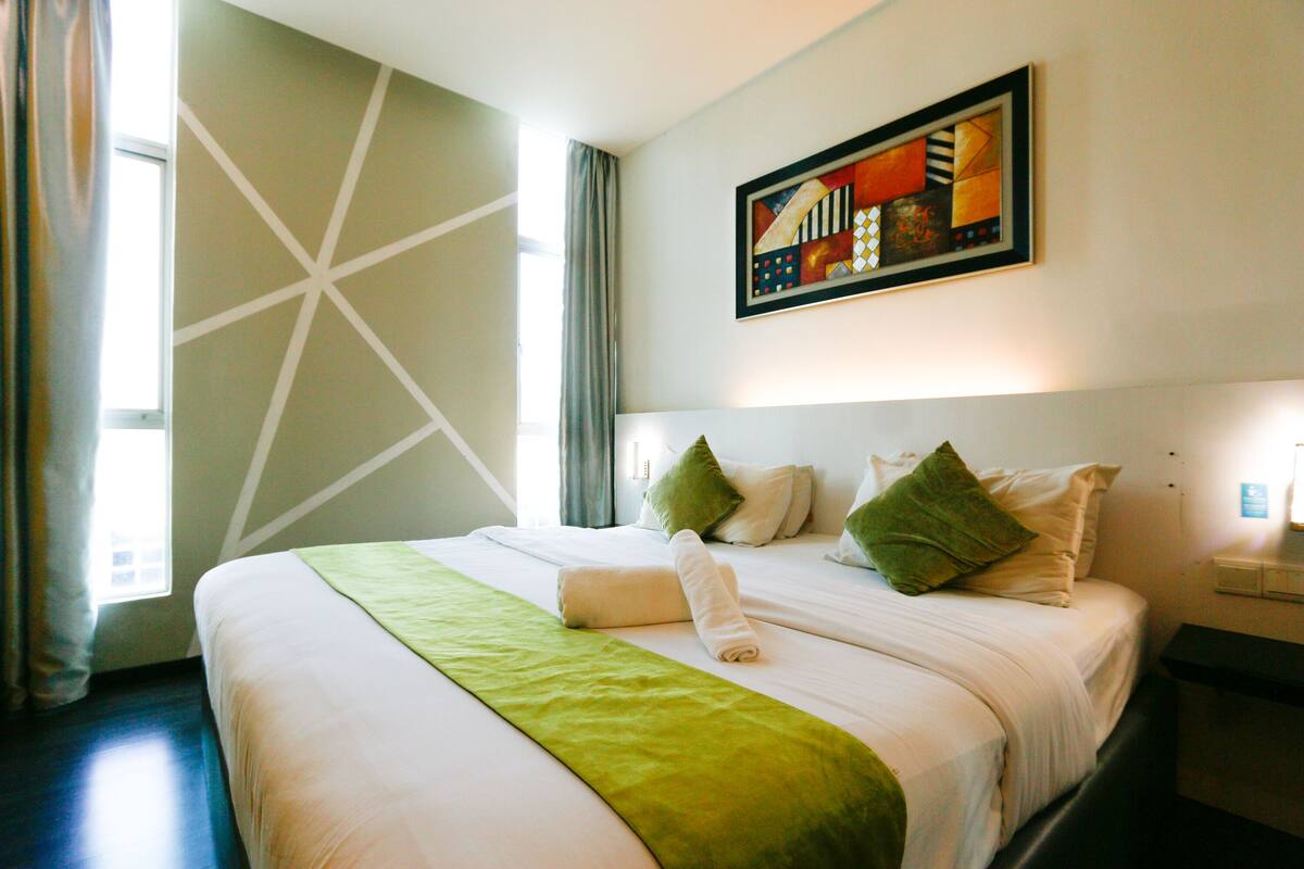 A hotel room with a large bed, a green throw blanket, and a painting on the wall.