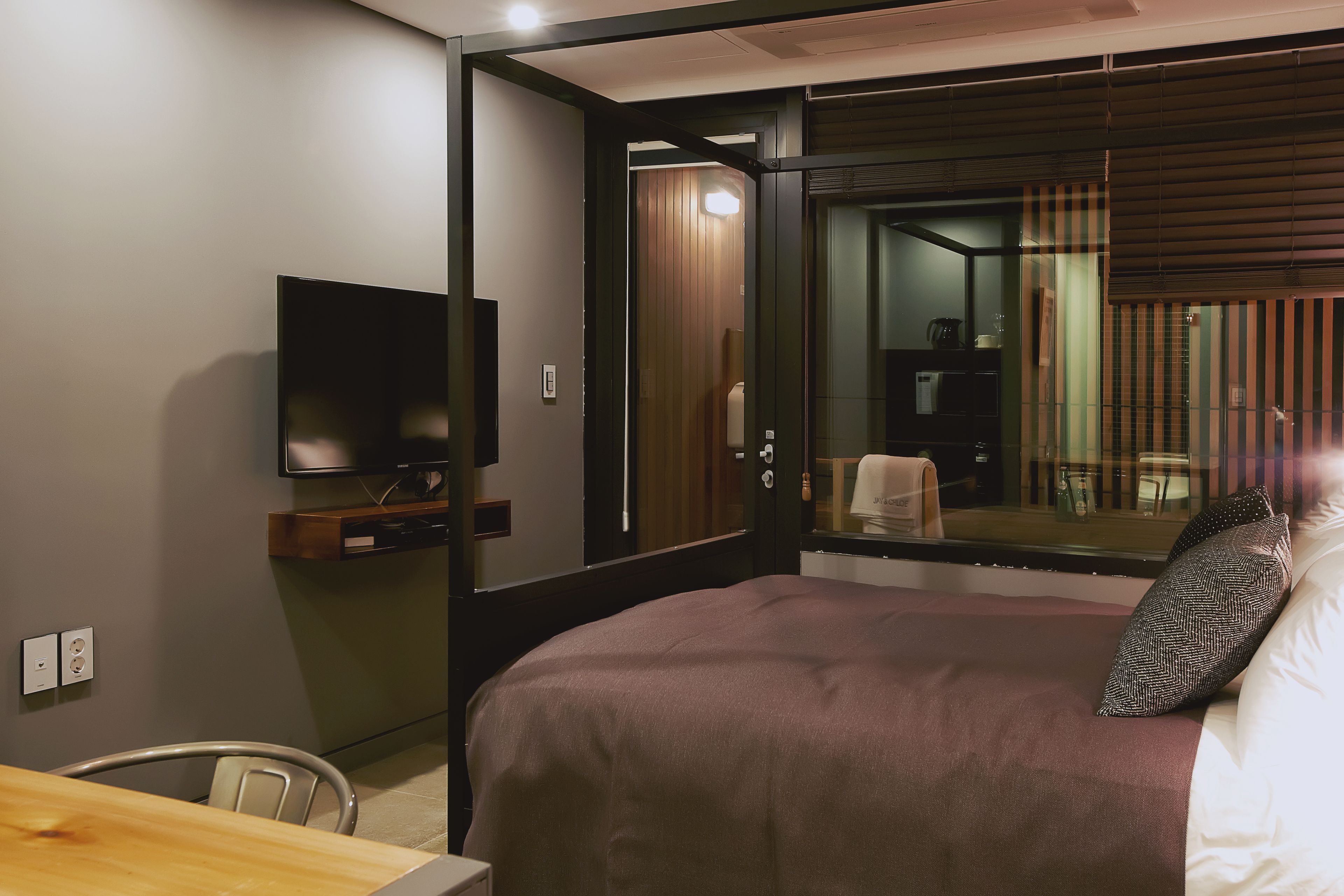A modern hotel room with a bed, a TV, a desk, and a window with blinds.
