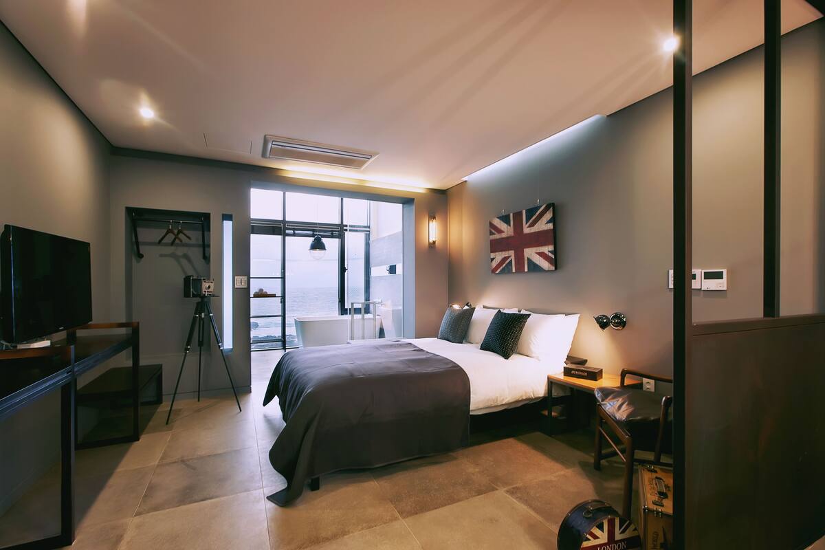 A modern hotel room with a large bed, a flat-screen TV, a tripod, and a view of the sea through the window.