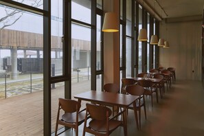 A spacious indoor area with large windows, wooden tables and chairs, and hanging pendant lights.