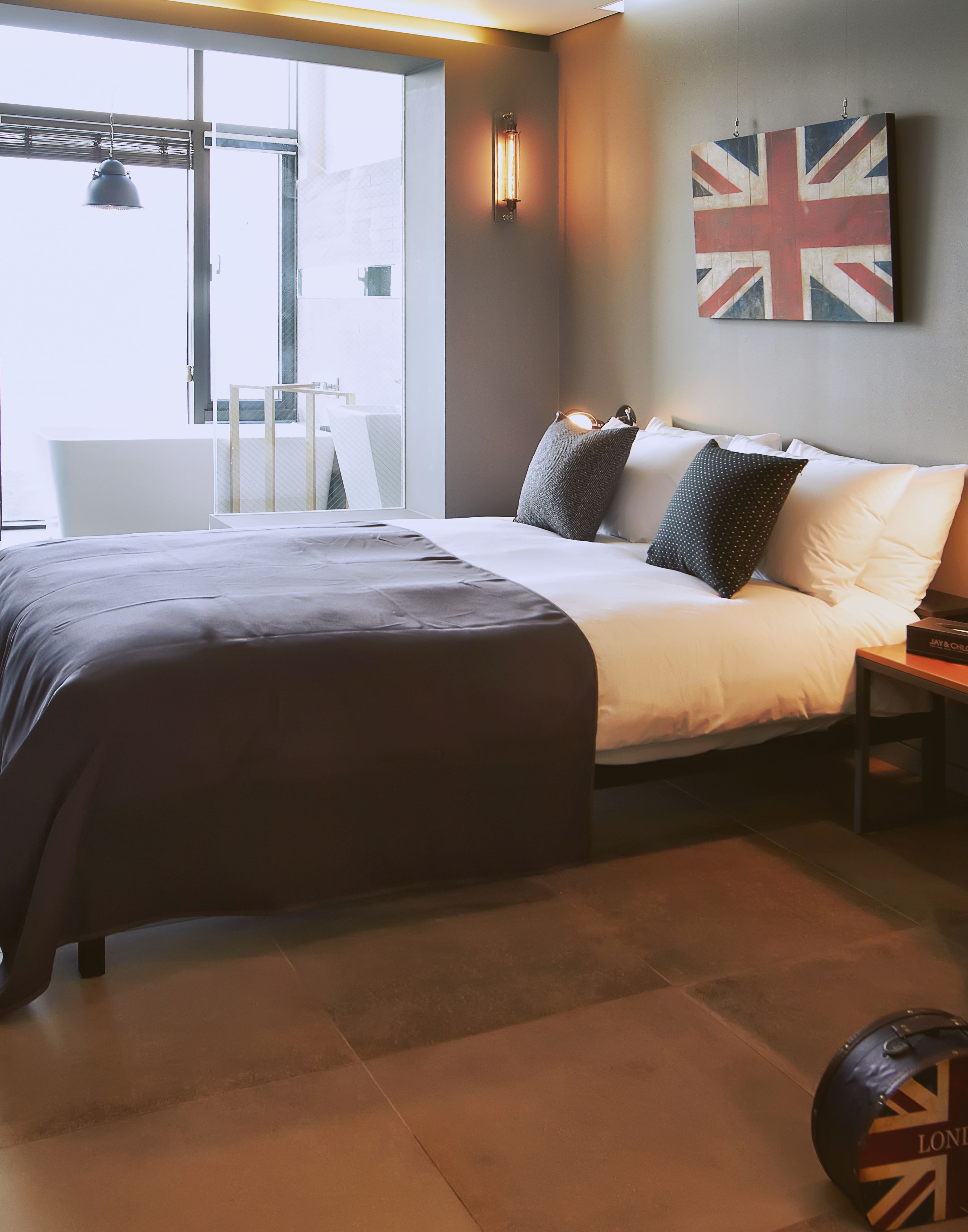 A modern hotel room with a large bed, a small bedside table, a wall-mounted light, and a window with a view.