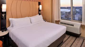 A hotel room with a bed, bedside lamps, a nightstand, a window with a city view, and an air conditioning unit.