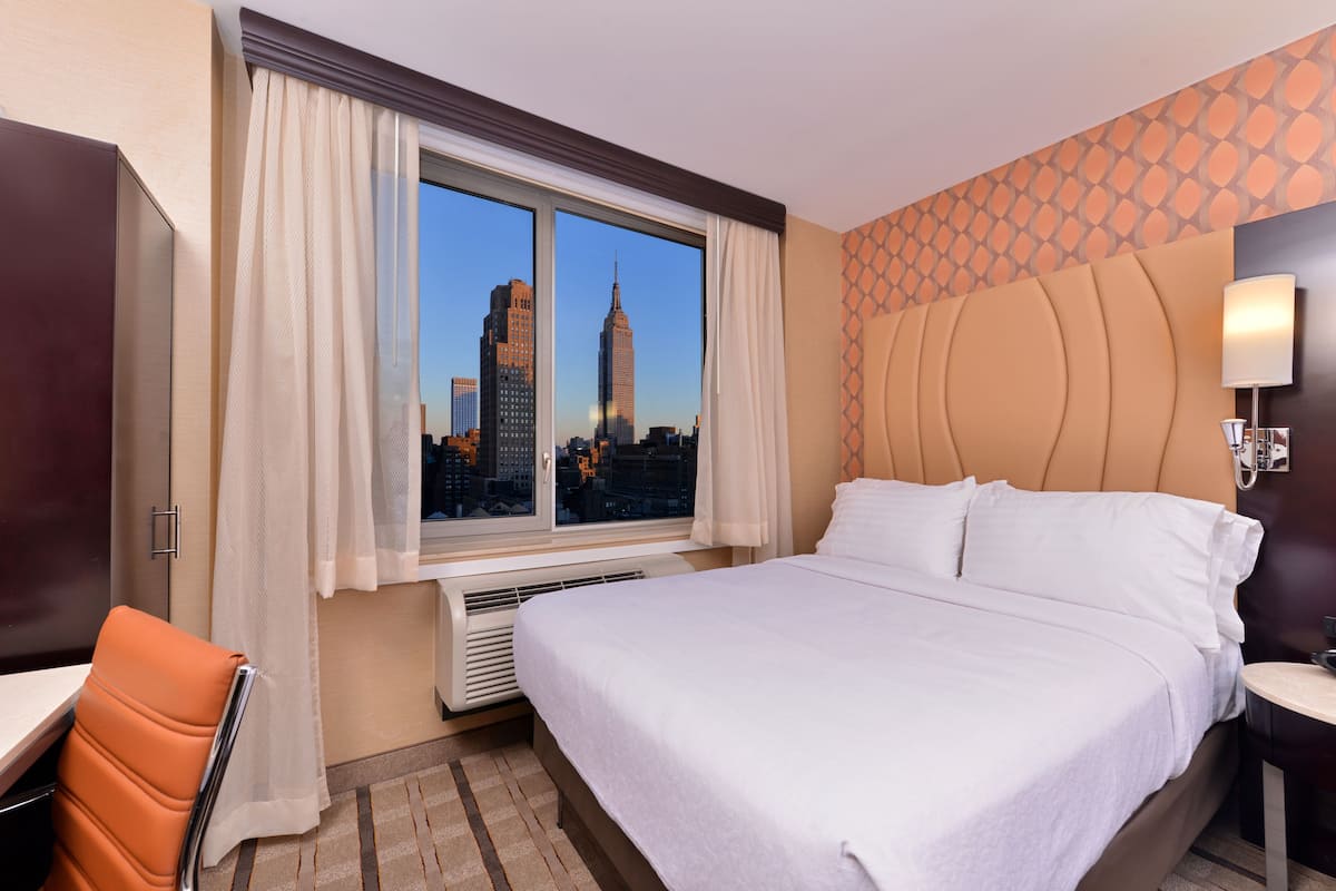 A hotel room with a large bed, a desk, and a chair. There is a window with a view of a city skyline.