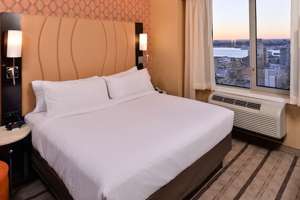 A hotel room with a large bed, a nightstand, a lamp, and a view of the city through a window. - Holiday Inn New York City - Times Square by IHG (New York)
