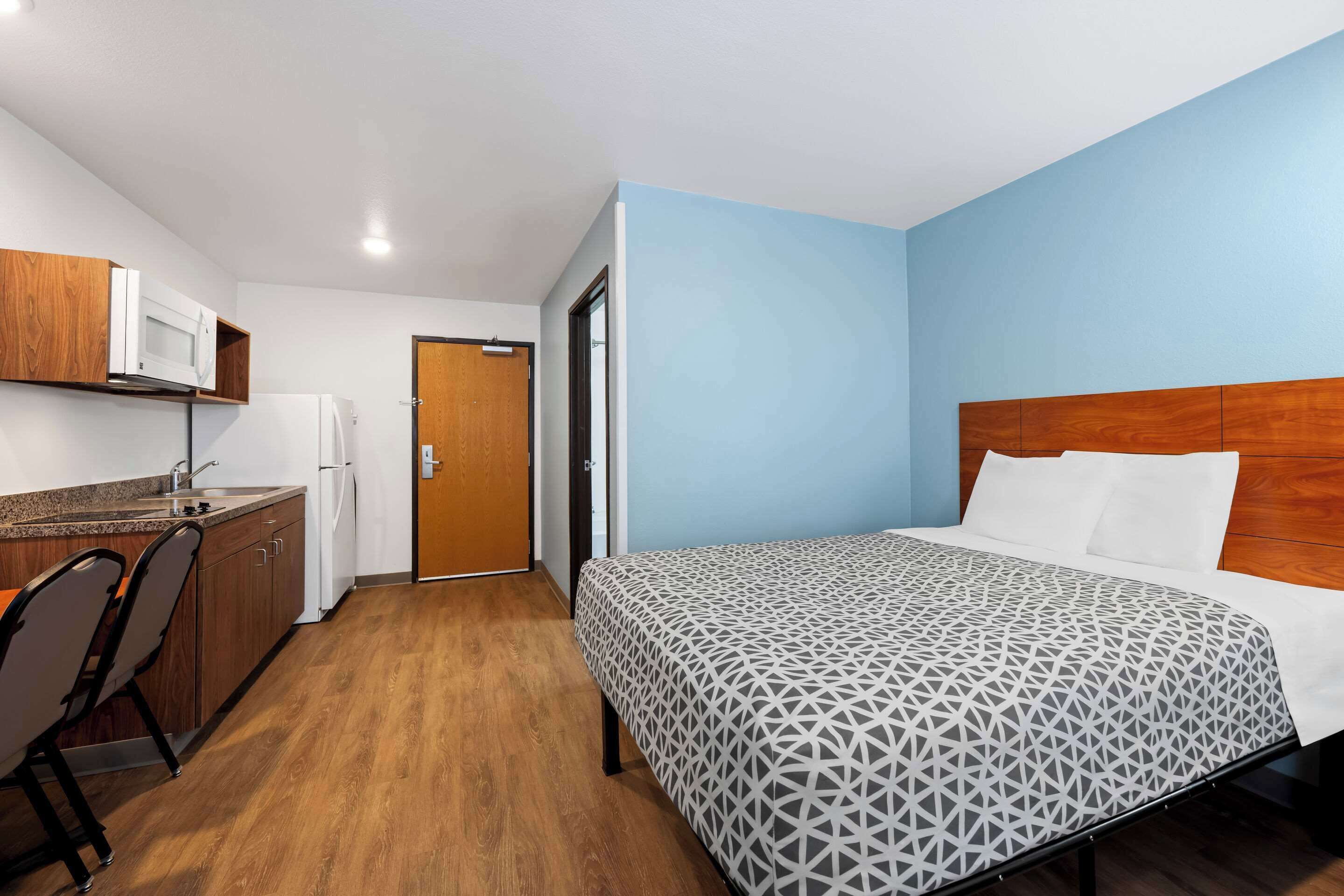 A compact hotel room with a bed, a kitchenette, and a wooden door.