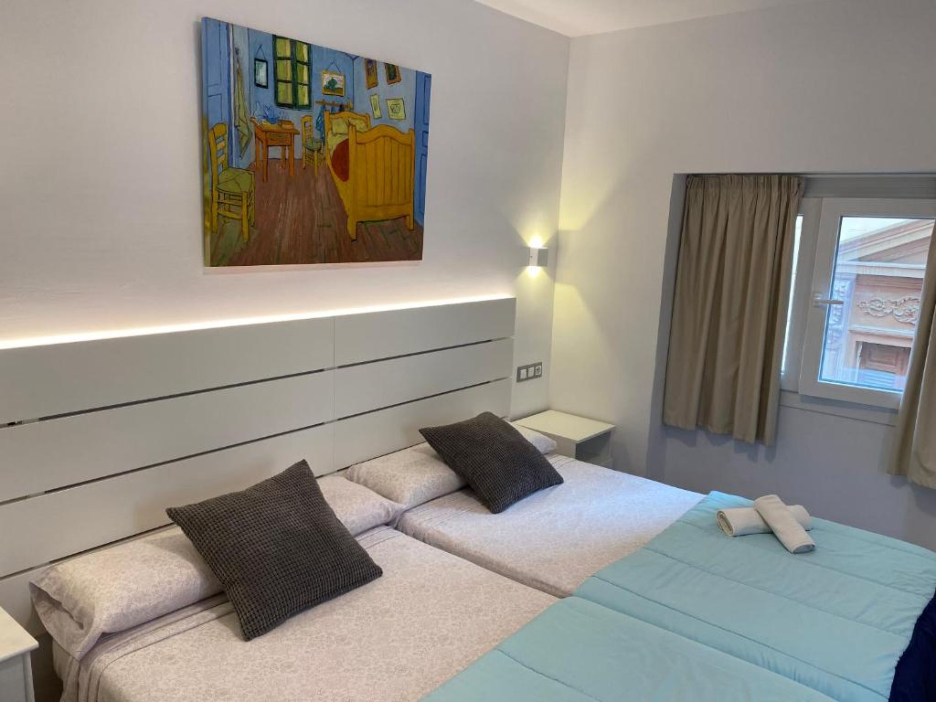 A double bed with a blue bedspread and two pillows, a painting above the bed, a bedside table with a lamp, and a window with curtains.