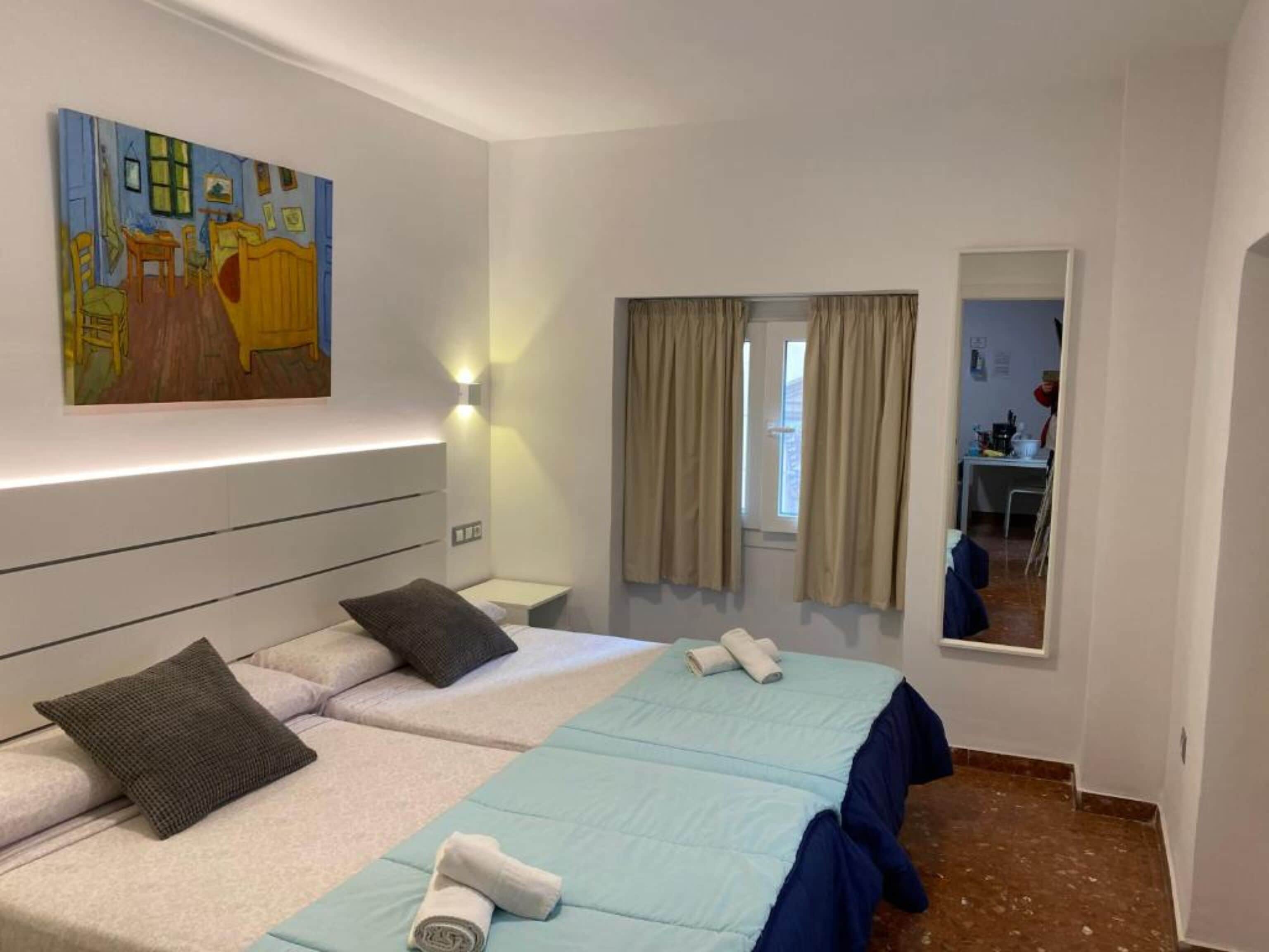 Habitación de hotel con dos camas, un cuadro en la pared, una ventana con cortinas y una pequeña mesa con una silla.