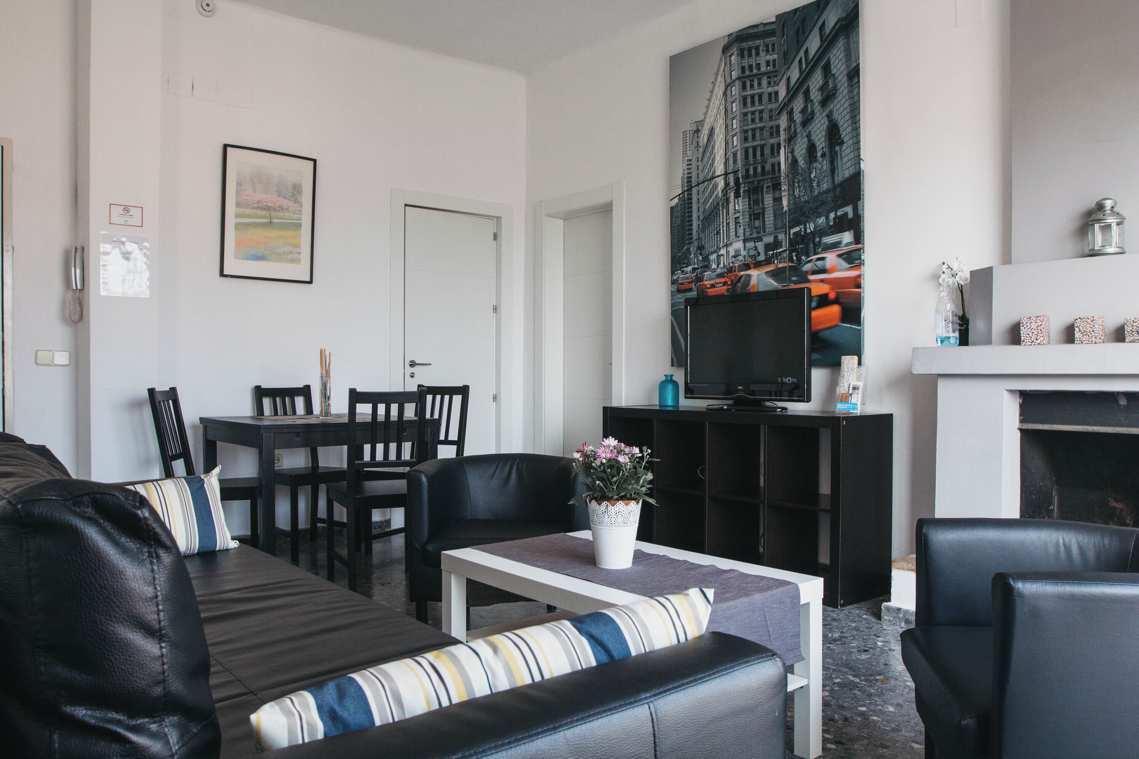 A modern living room with a black leather sofa, a dining table with chairs, a TV on a stand, and a fireplace.