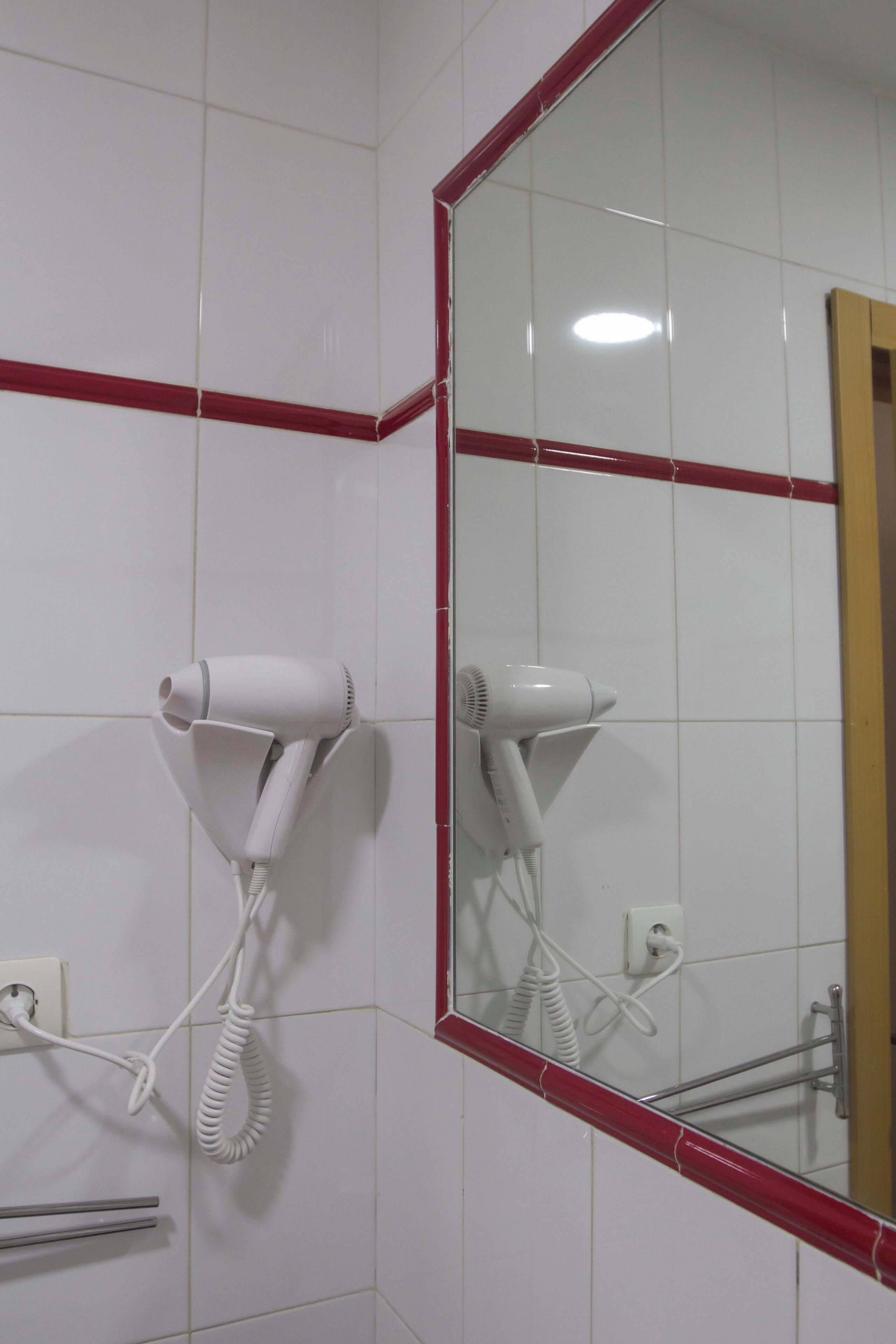 Un baño con azulejos blancos, una zona de ducha con baldosas rojas y un secador de pelo montado en la pared.