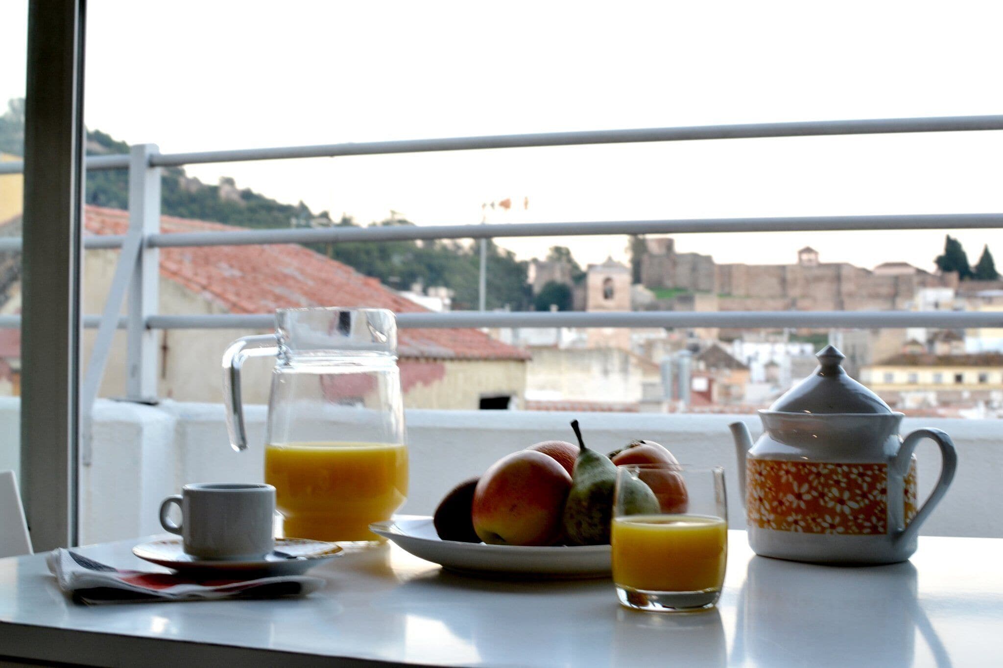A breakfast setup with a pitcher of orange juice, a teapot, a plate of fruit, and a cup on a table by a window overlooking a cityscape.
