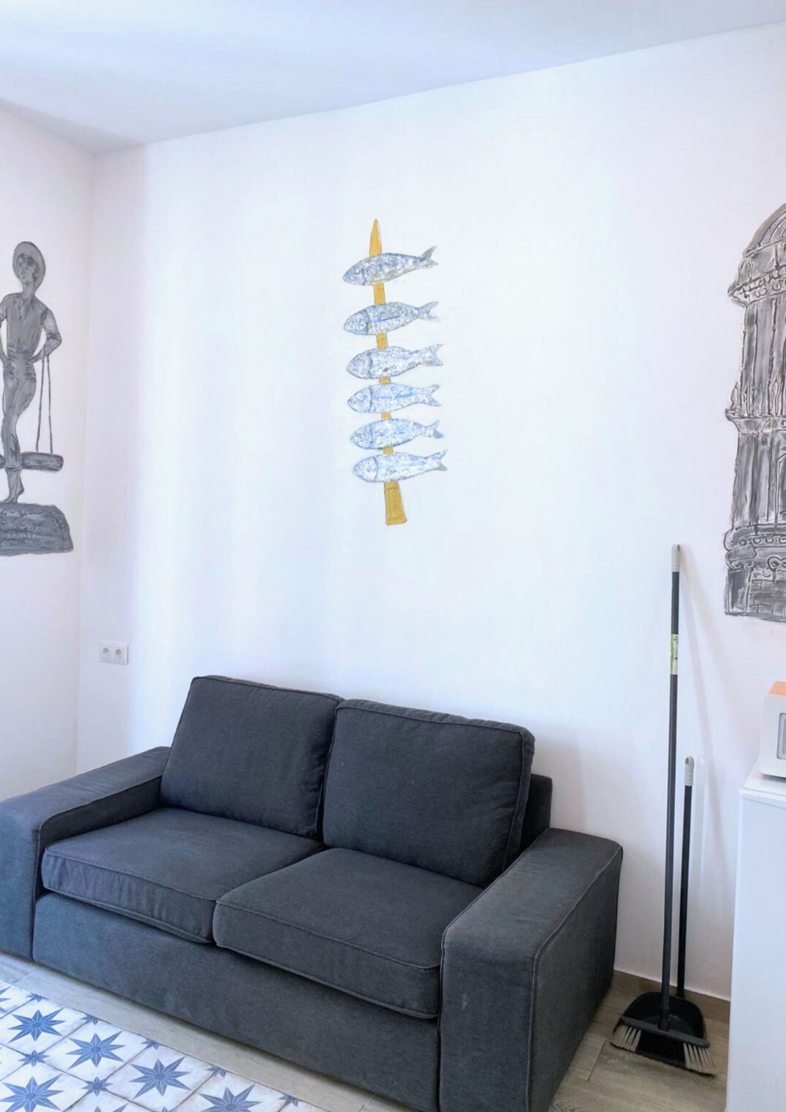 Ein Wohnzimmer mit einem grauen Sofa, einer Wand mit einem Fischgemälde, einem Besen und einem kleinen Tisch mit einer Lampe.