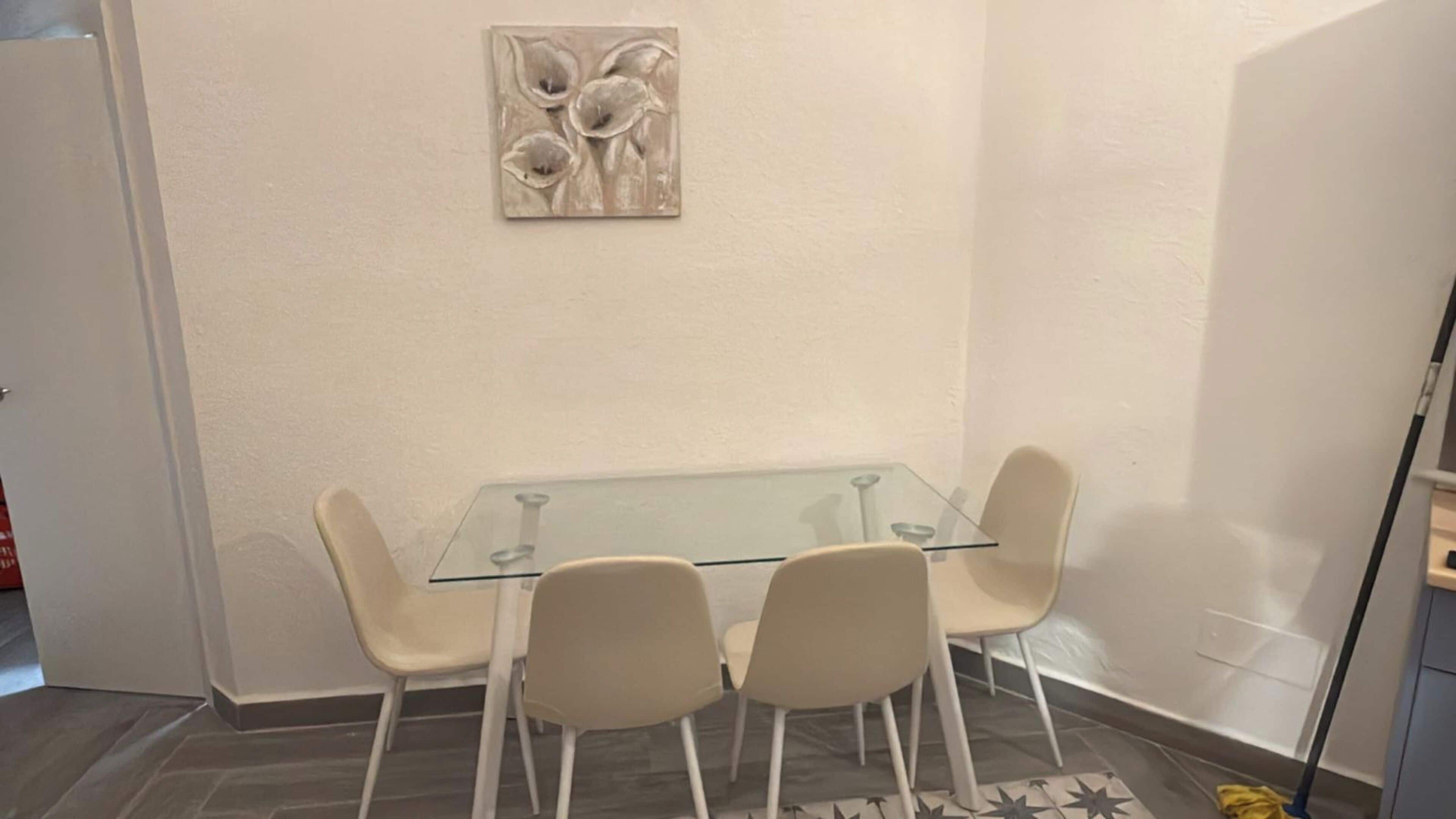 A dining area with a glass table and four chairs, a wall-mounted artwork, and a door on the left.