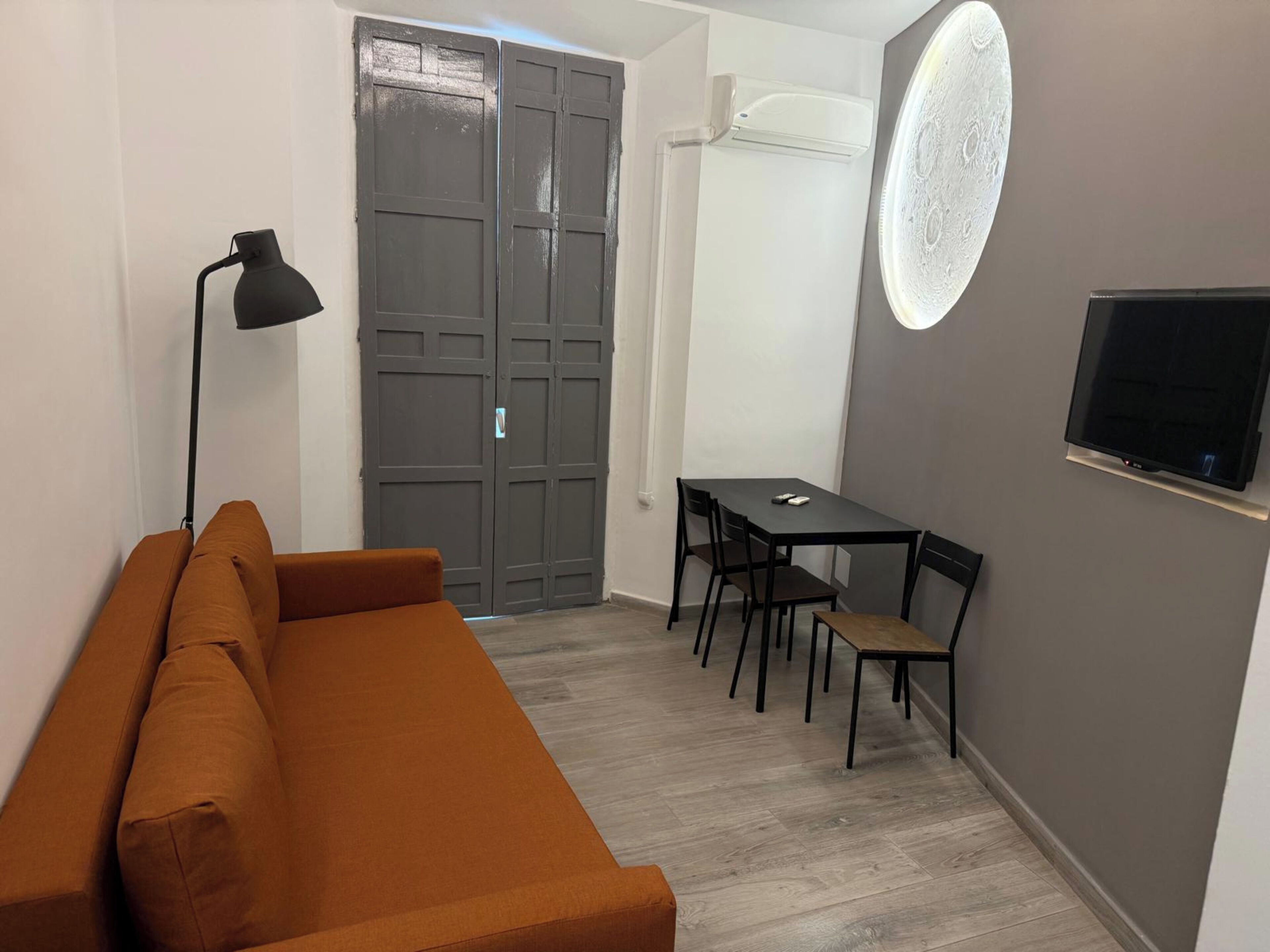 A compact living space with a sofa, a dining table with chairs, a TV mounted on the wall, and a door with an air conditioning unit.