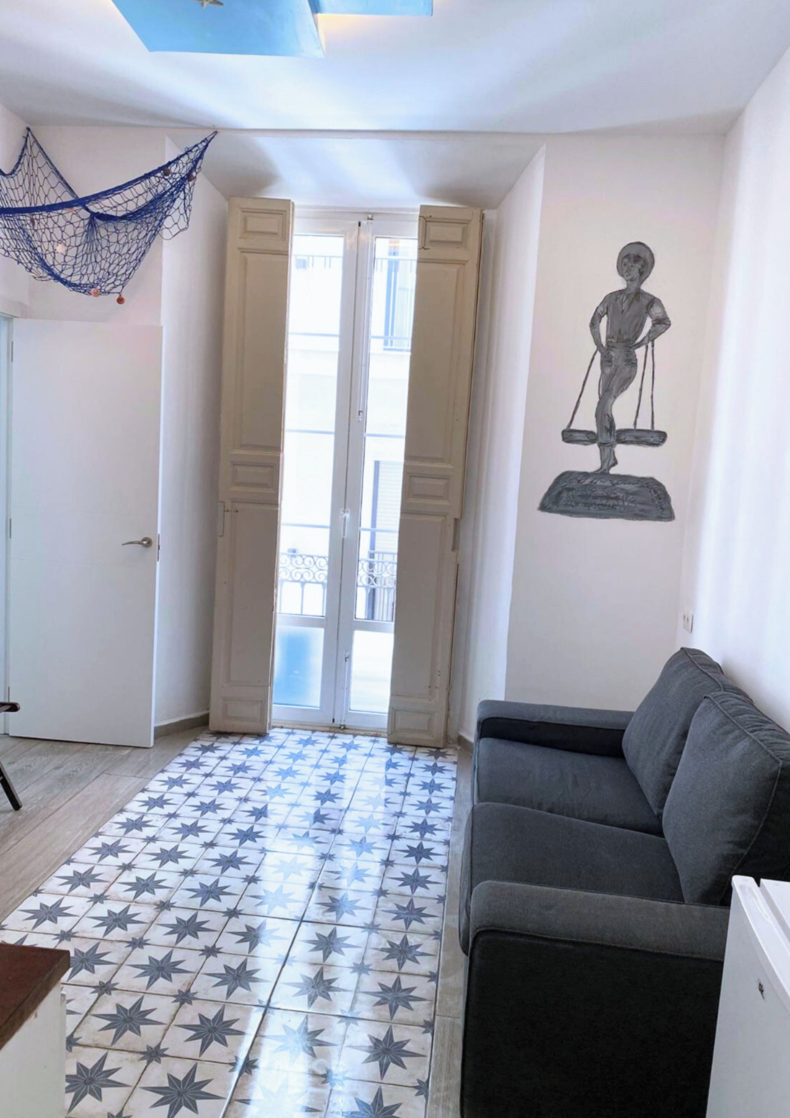 Une pièce avec un canapé gris, un sol carrelé orné de motifs en étoile, une porte avec des panneaux de verre et un mur sur lequel est accrochée une sculpture en forme d’échelle.