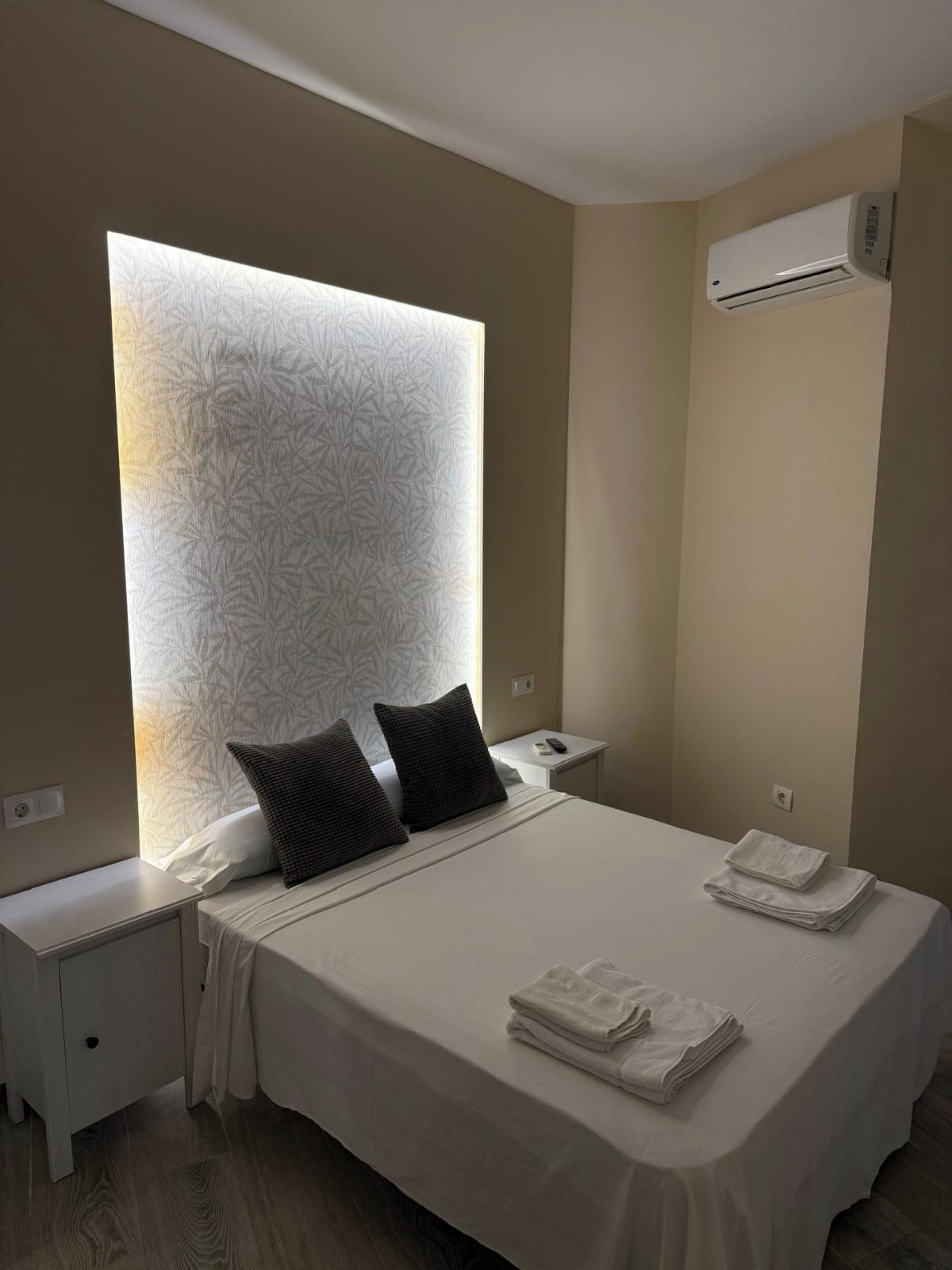 A modern hotel room with a large bed, two bedside tables, a wall-mounted air conditioner, and a patterned headboard.