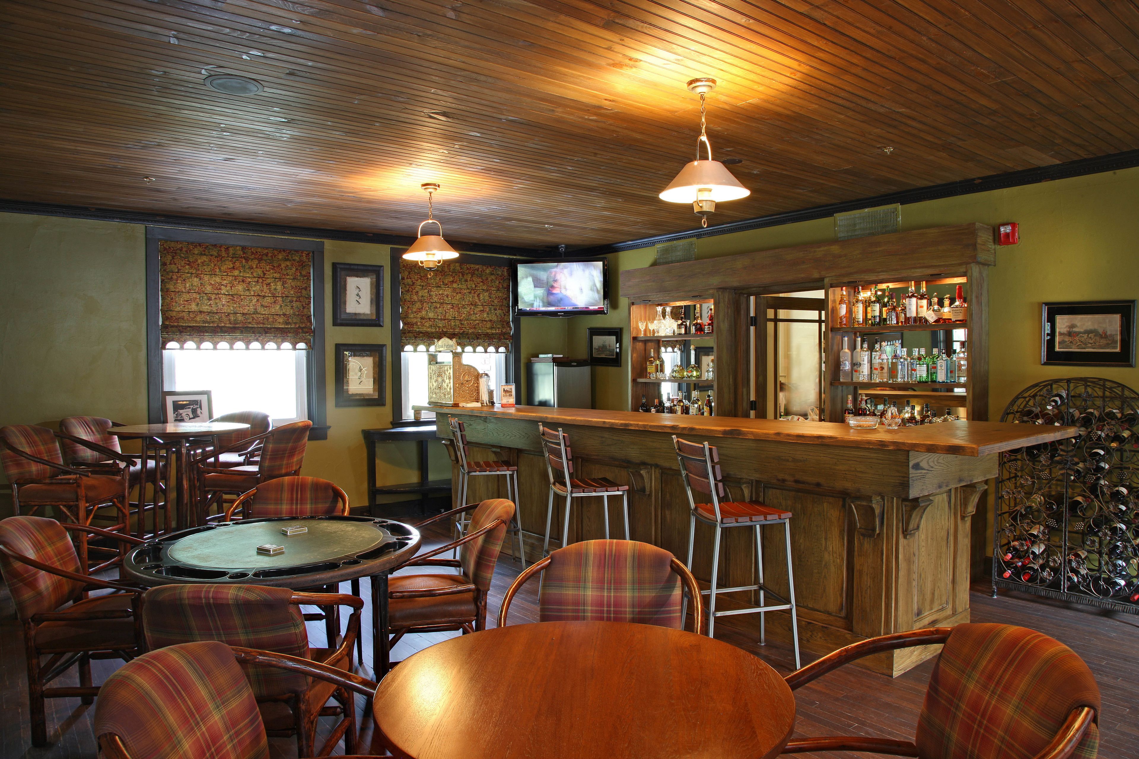 A bar area with wooden ceiling, plaid chairs, a billiards table, and a bar counter with bottles.