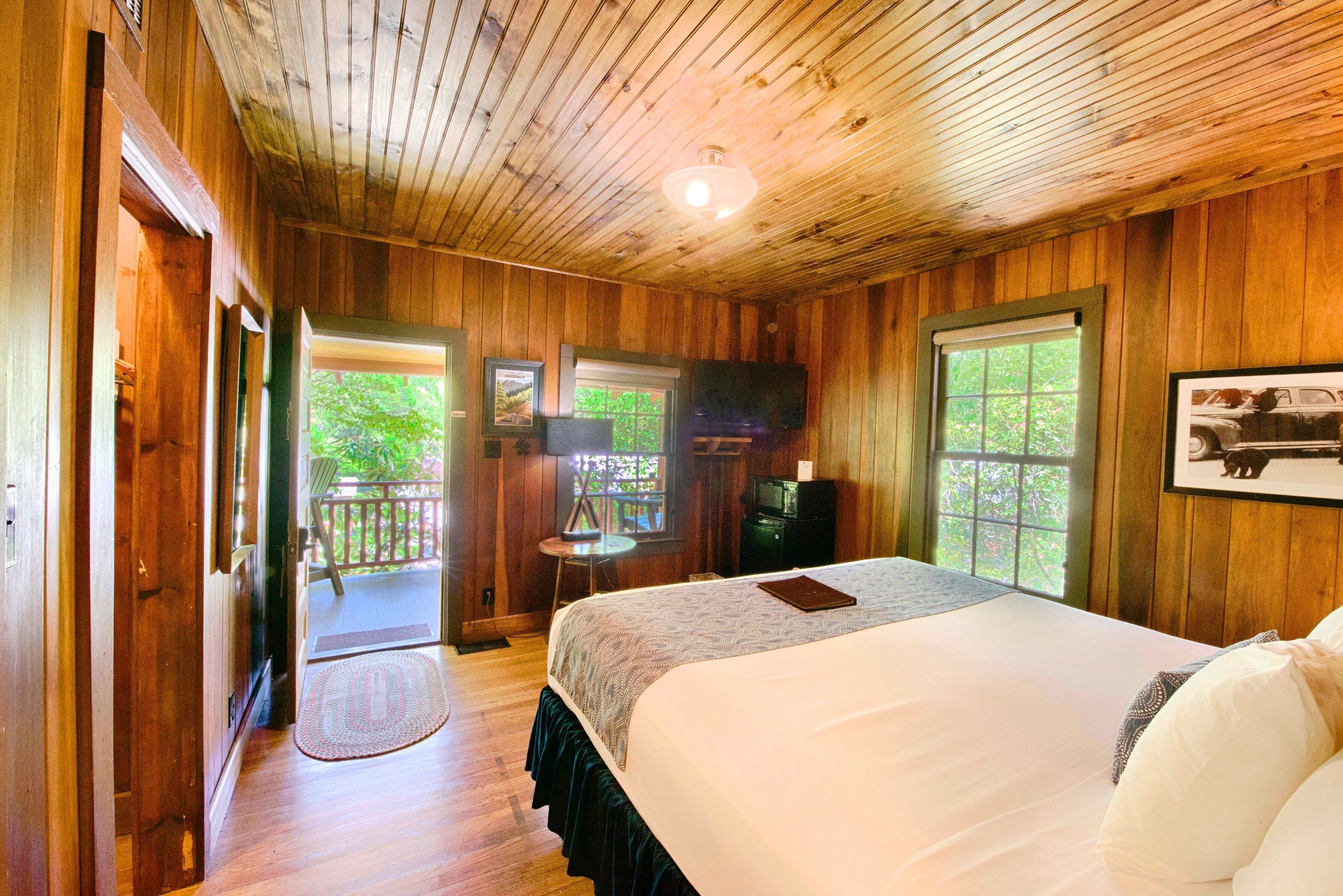 A wooden cabin bedroom with a bed, a television, and a window with a view of greenery.