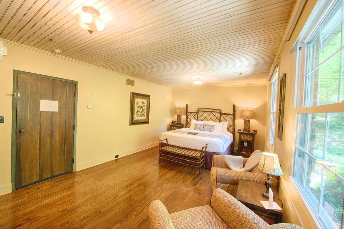 A bedroom with a wooden floor, a bed with a headboard, a bench, a chair, a nightstand with a lamp, and a window with a view of greenery.