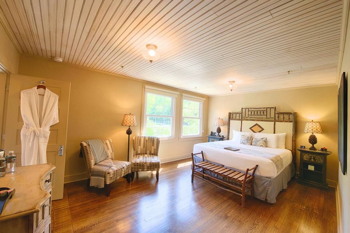 A bedroom with a wooden ceiling, a bed with a headboard, two chairs, a bench, and a nightstand with a lamp.