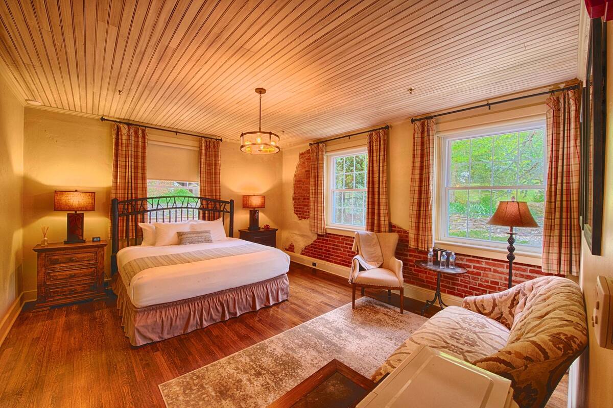 A bedroom with a wooden ceiling, a large bed, a chair, a nightstand, a lamp, and a window with curtains.