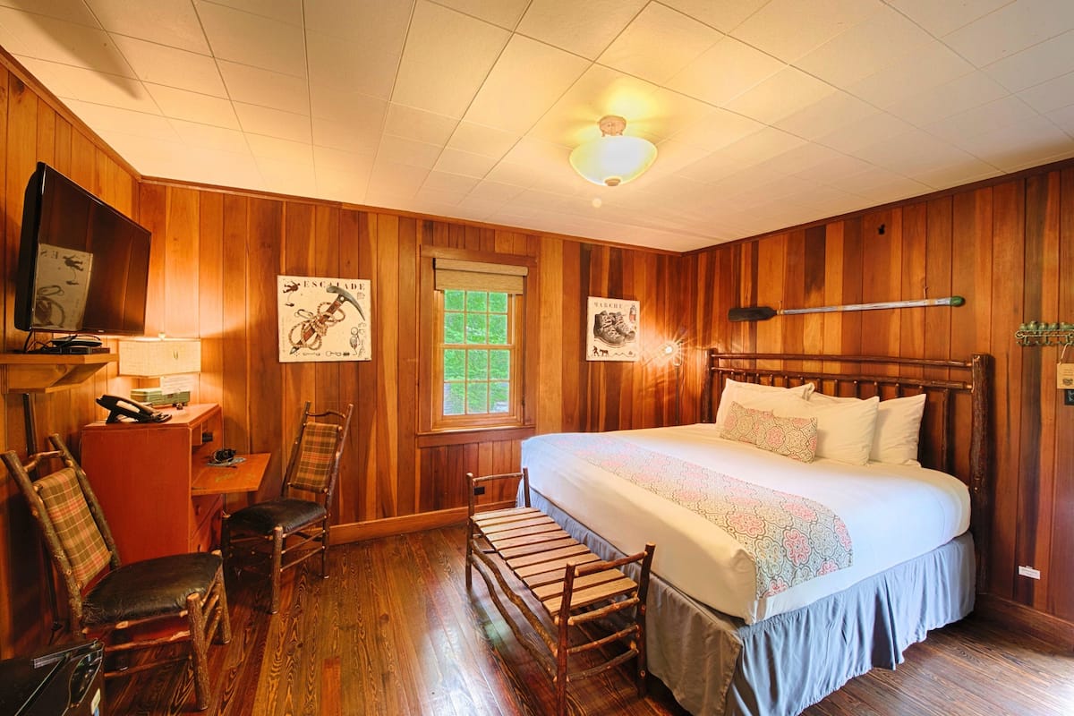 A wooden-paneled room with a bed, two chairs, a desk, and a television.