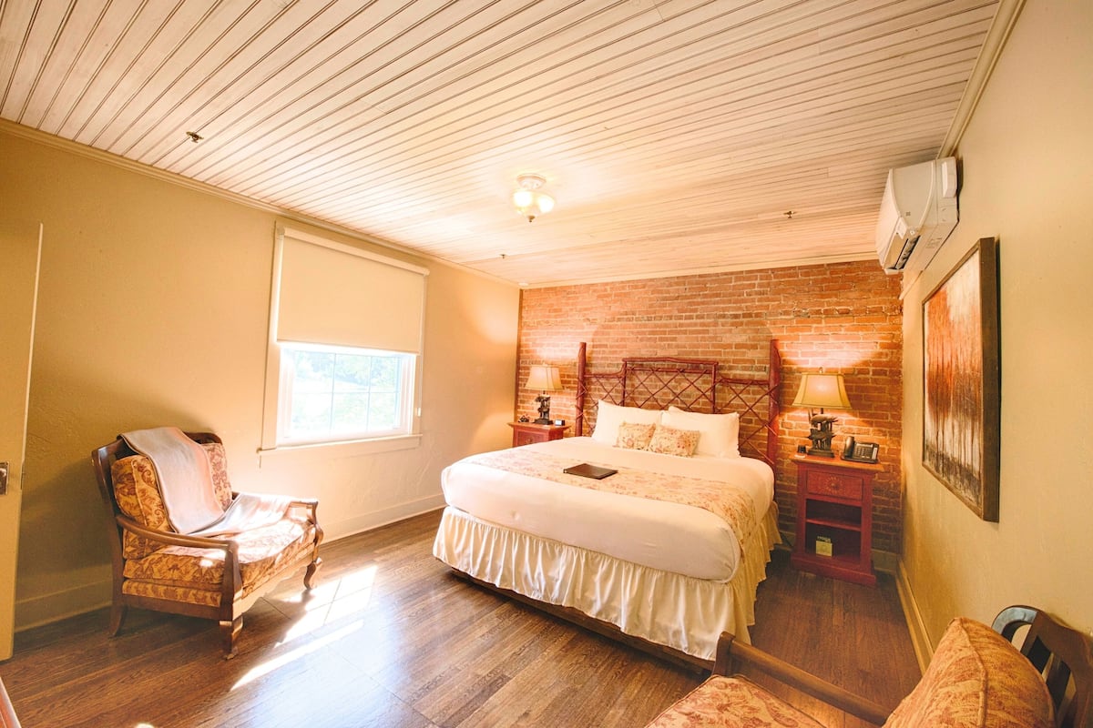 A bedroom with a large bed, a wooden chair, a nightstand with a lamp, and a brick wall.