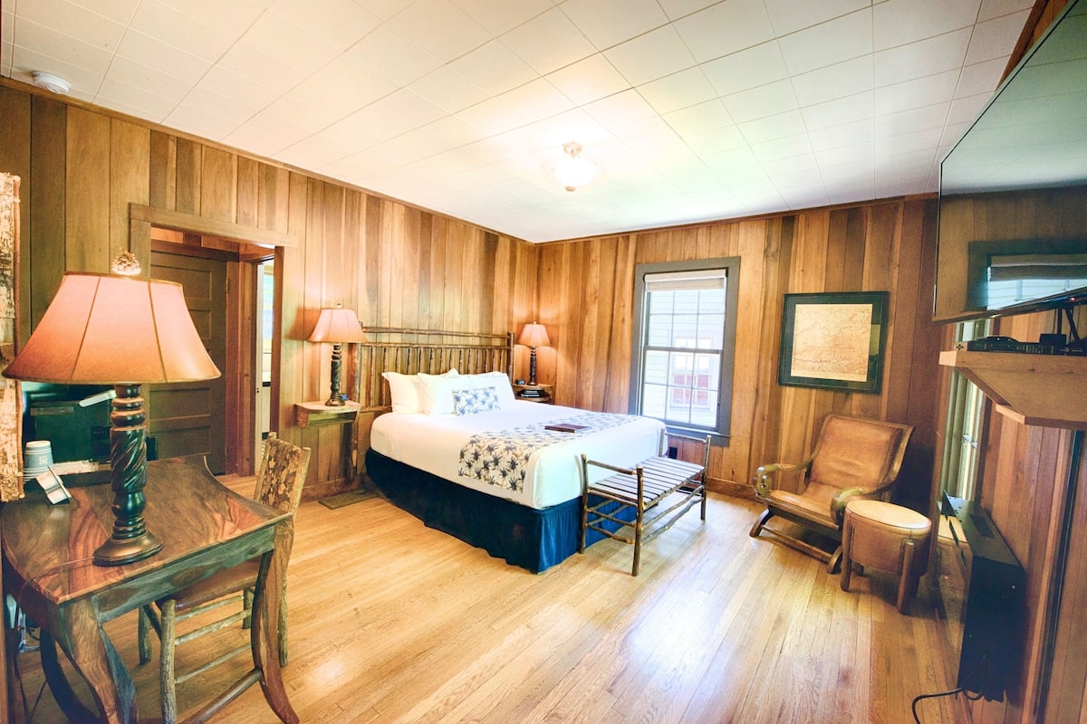A wooden-paneled room with a bed, two bedside lamps, a chair, and a table.