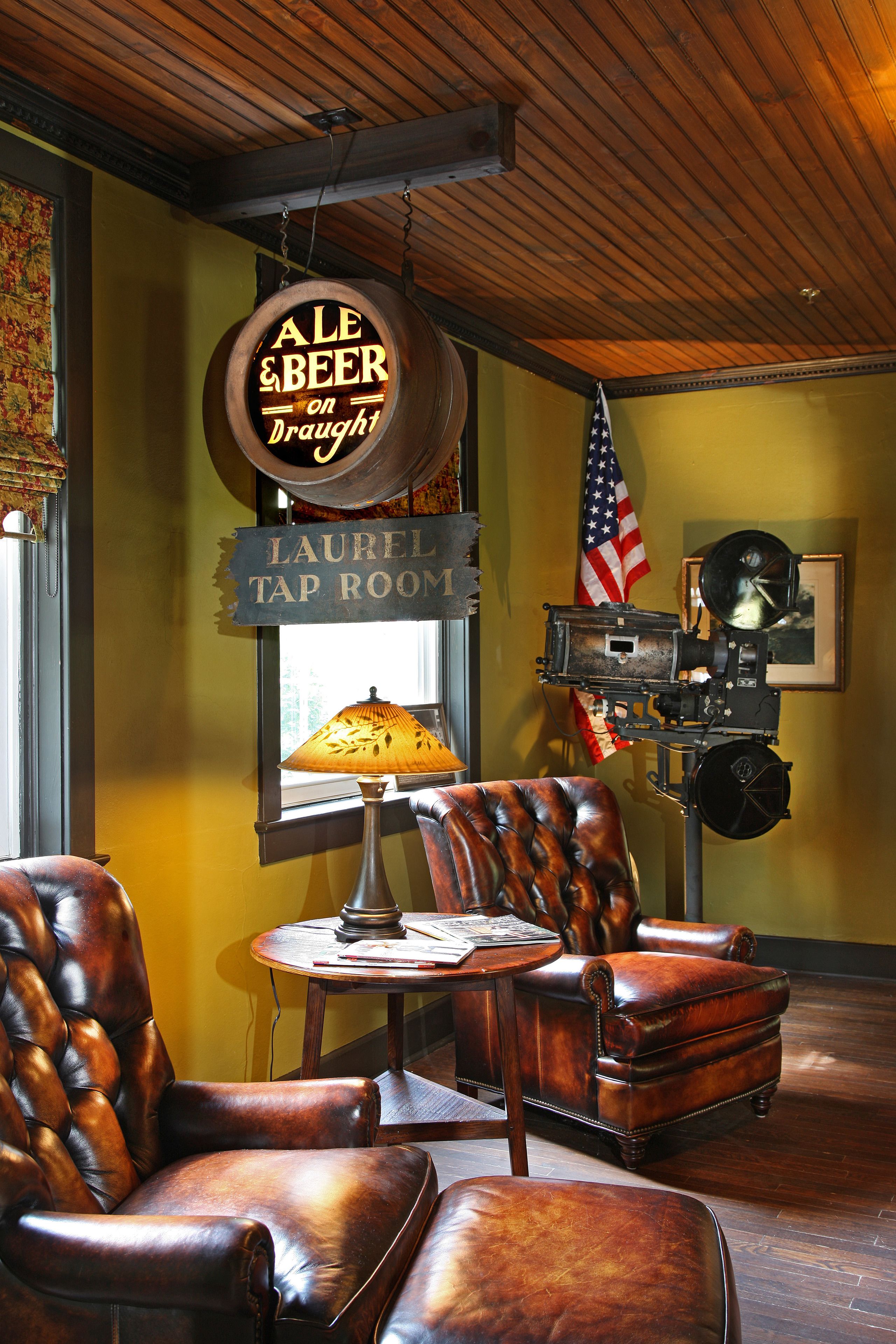 A tap room with leather chairs, a table, an American flag, and a sign reading "ALE & BEER on Draught."