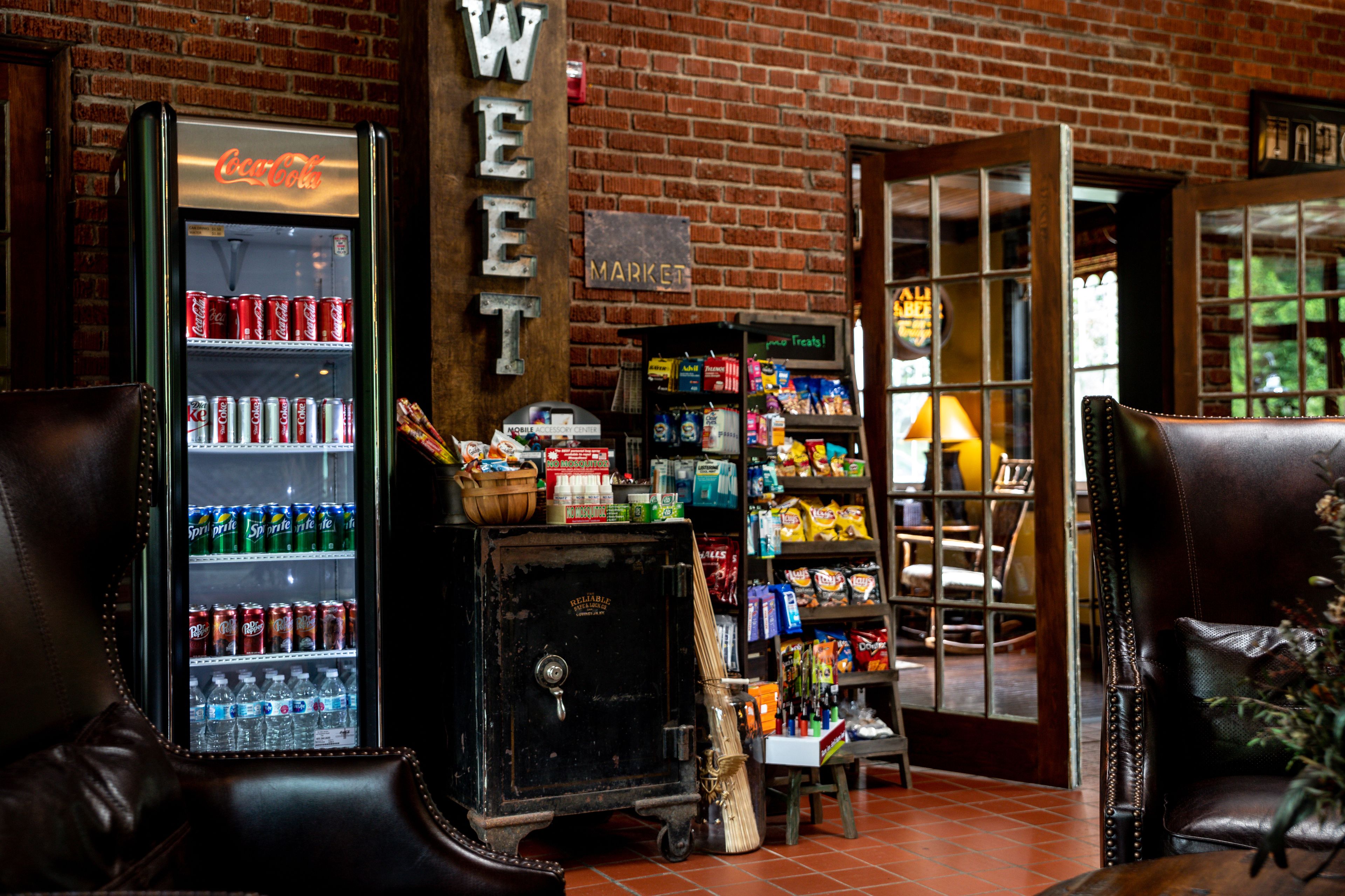 A vintage Coca-Cola refrigerator, a broom, and shelves stocked with snacks and beverages in a rustic brick building.