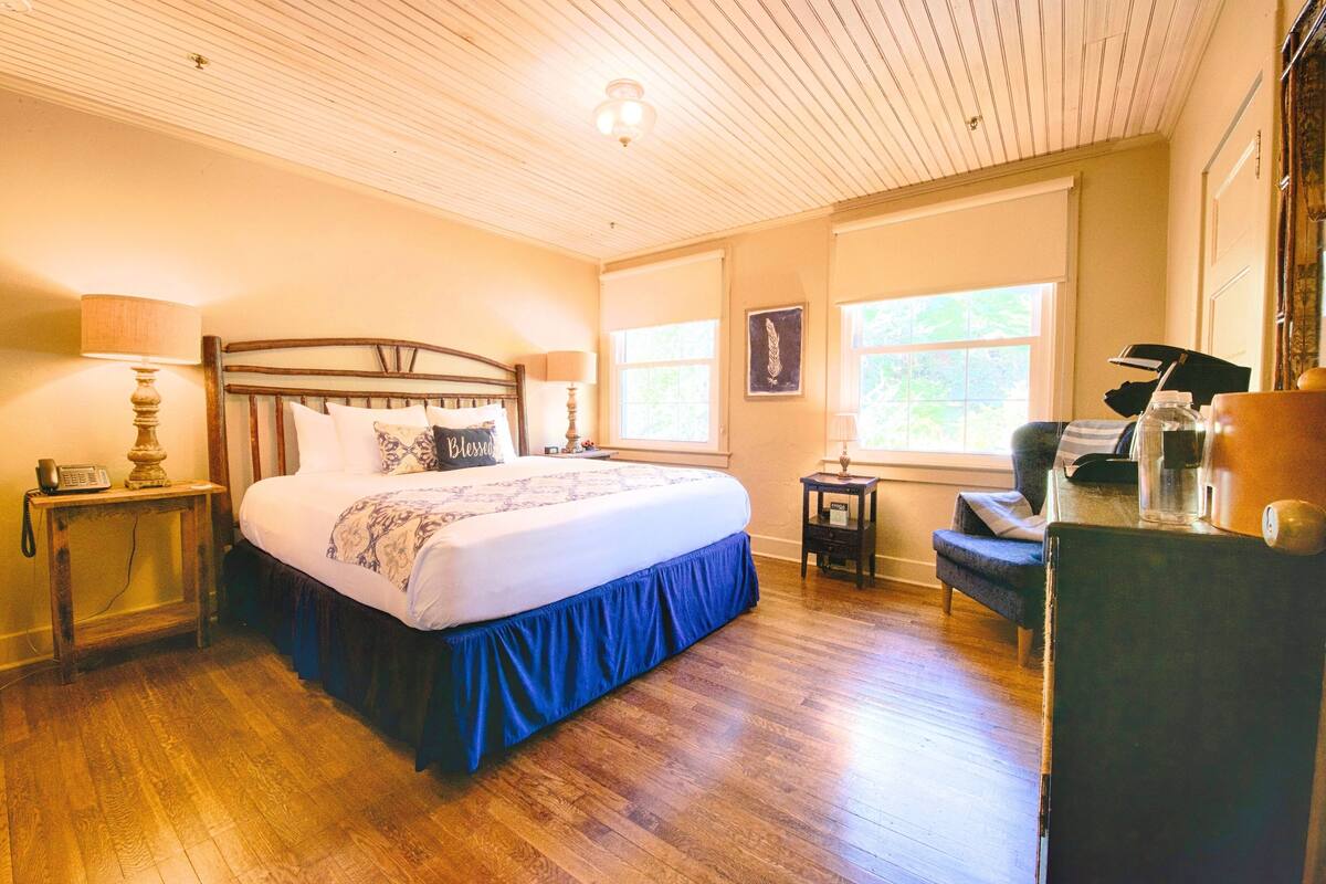 A bedroom with a wooden floor, a bed with a blue skirt, two bedside tables with lamps, a chair, and a window with a view of greenery.