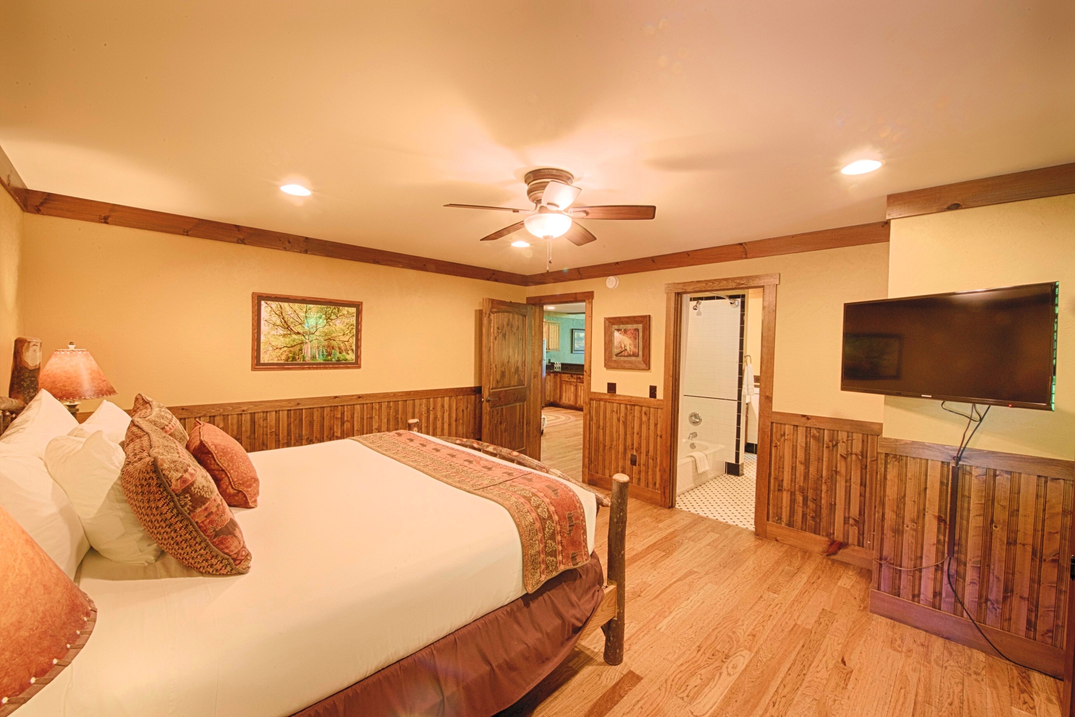 A bedroom with a wooden bed, a ceiling fan, a TV mounted on the wall, and a wooden floor.