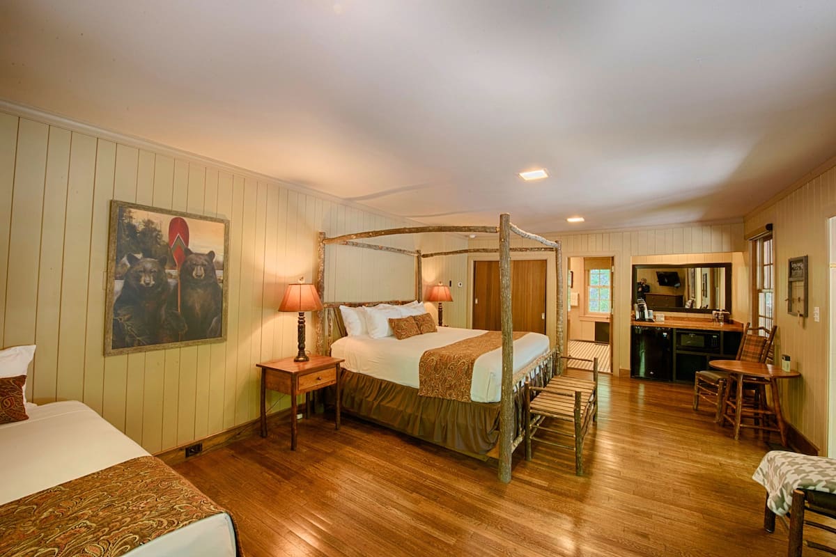 A four-poster bed with a canopy, a painting on the wall, a wooden floor, a kitchen area, and a dining table with chairs.