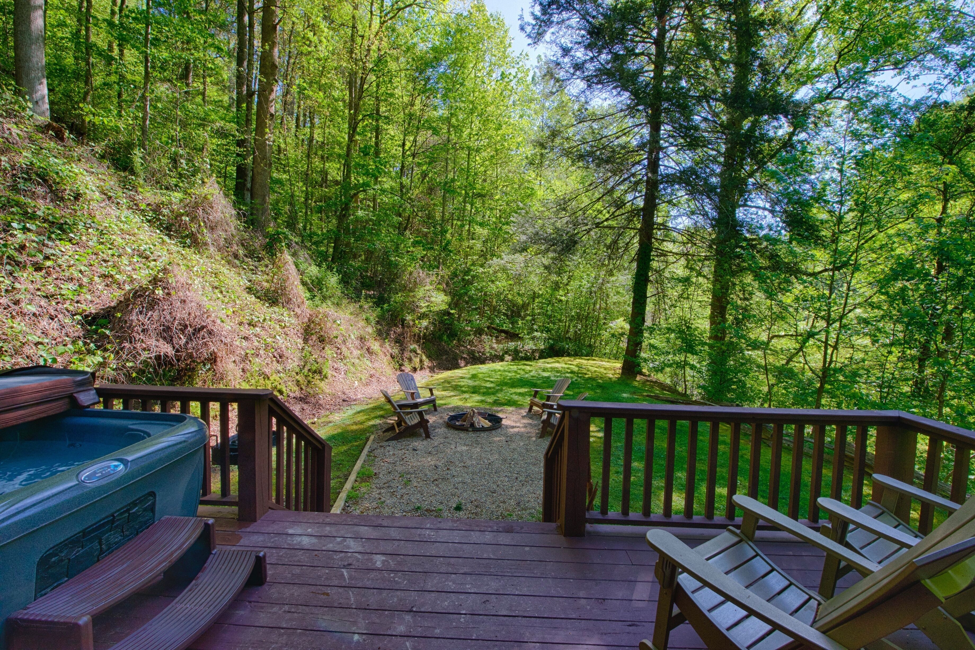 A deck with Adirondack chairs and a hot tub overlooking a forested area with a fire pit.