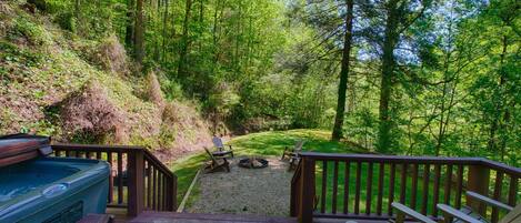 A deck with Adirondack chairs and a hot tub overlooking a forested area with a fire pit.