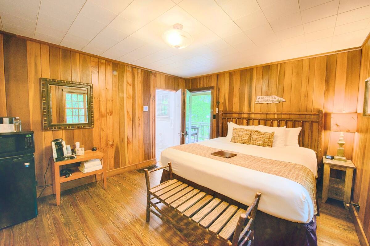 A wooden-paneled bedroom with a bed, a nightstand, a lamp, a mirror, a door, and a small table with a coffee maker and cups.