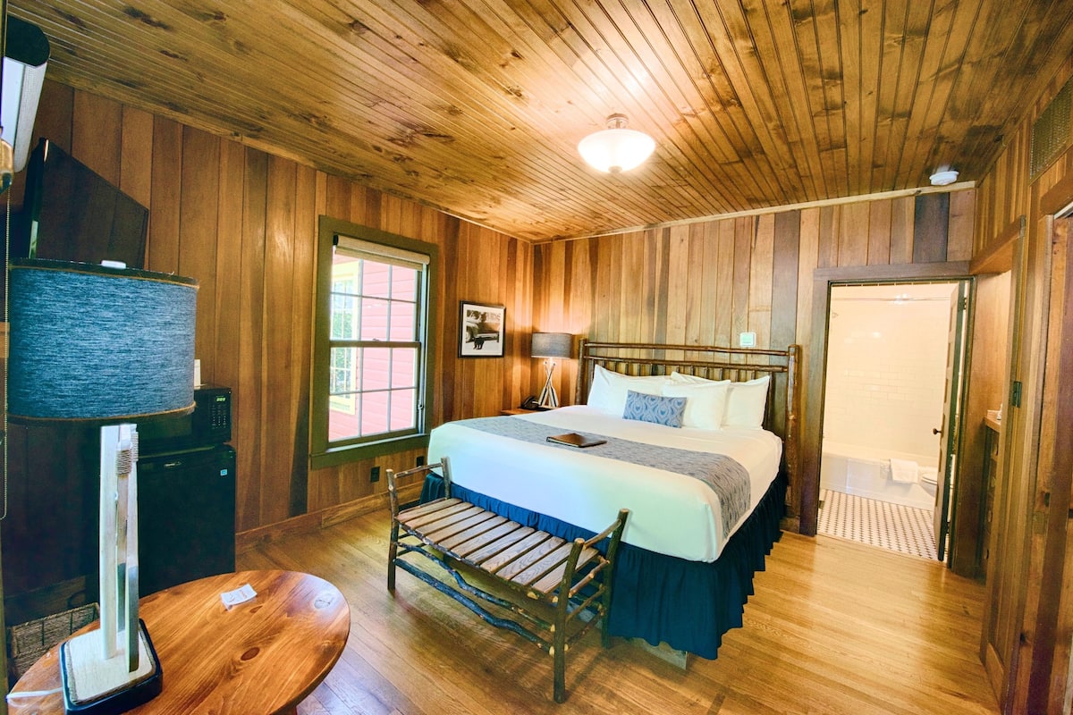 A wooden-paneled bedroom with a bed, bedside table, lamp, and a bench.