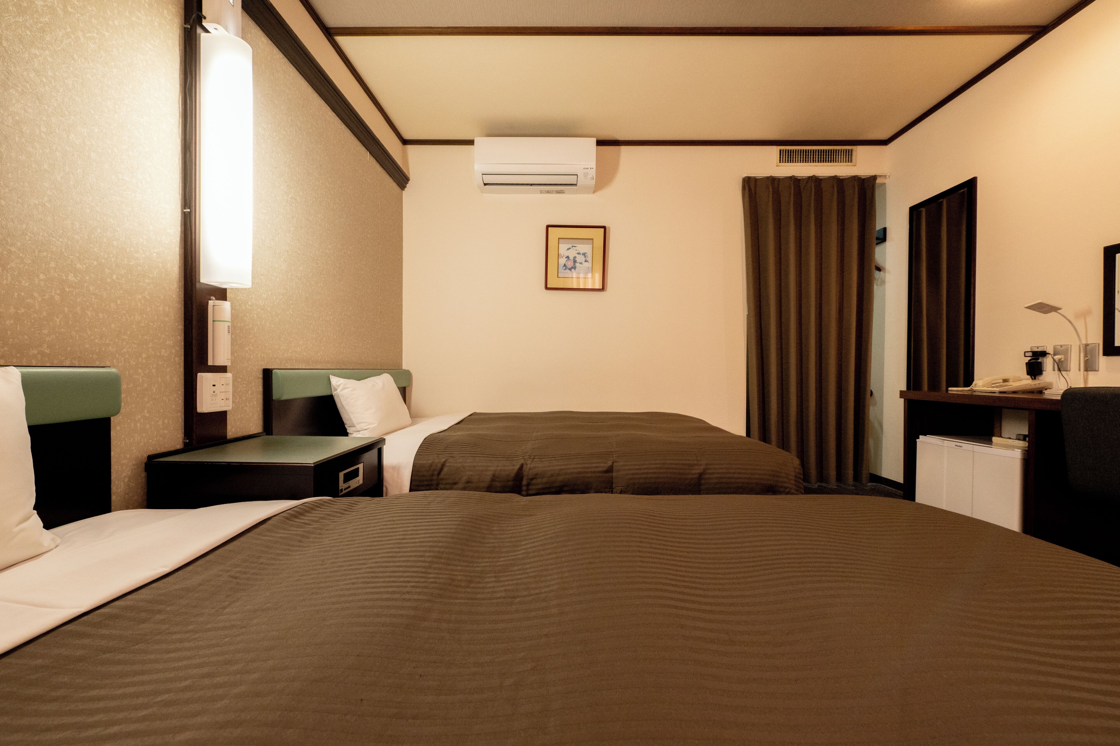 Habitación de hotel con dos camas, un escritorio, una silla, un equipo de aire acondicionado, un cuadro en la pared y una ventana con cortinas.