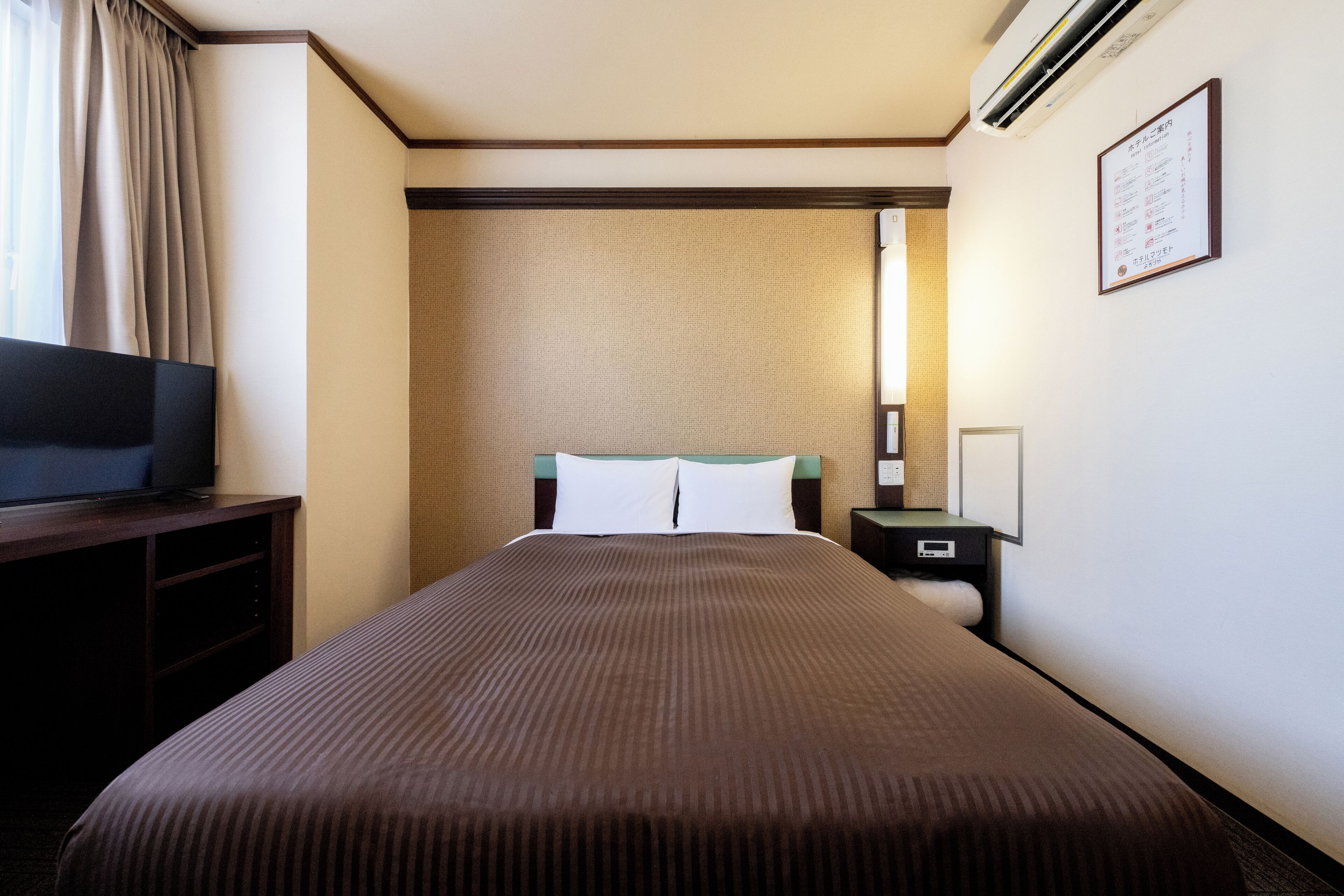 A hotel room with a bed, a television on a stand, a nightstand, and a wall-mounted air conditioner.