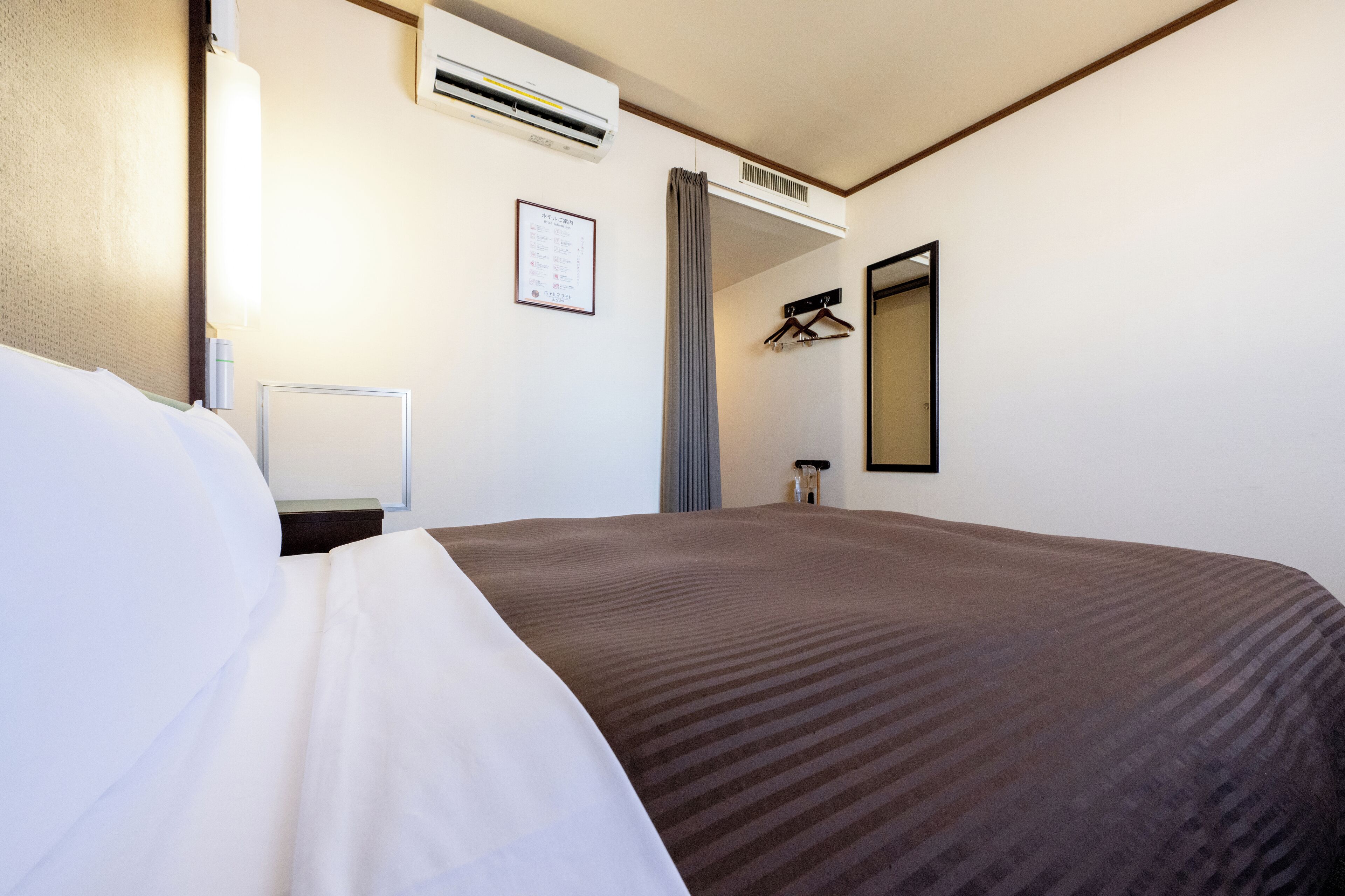 Ein Hotelzimmer mit einem großen Bett, einem Nachttisch, einer Klimaanlage, einem Spiegel und einem an der Wand befestigten Kleiderhaken.