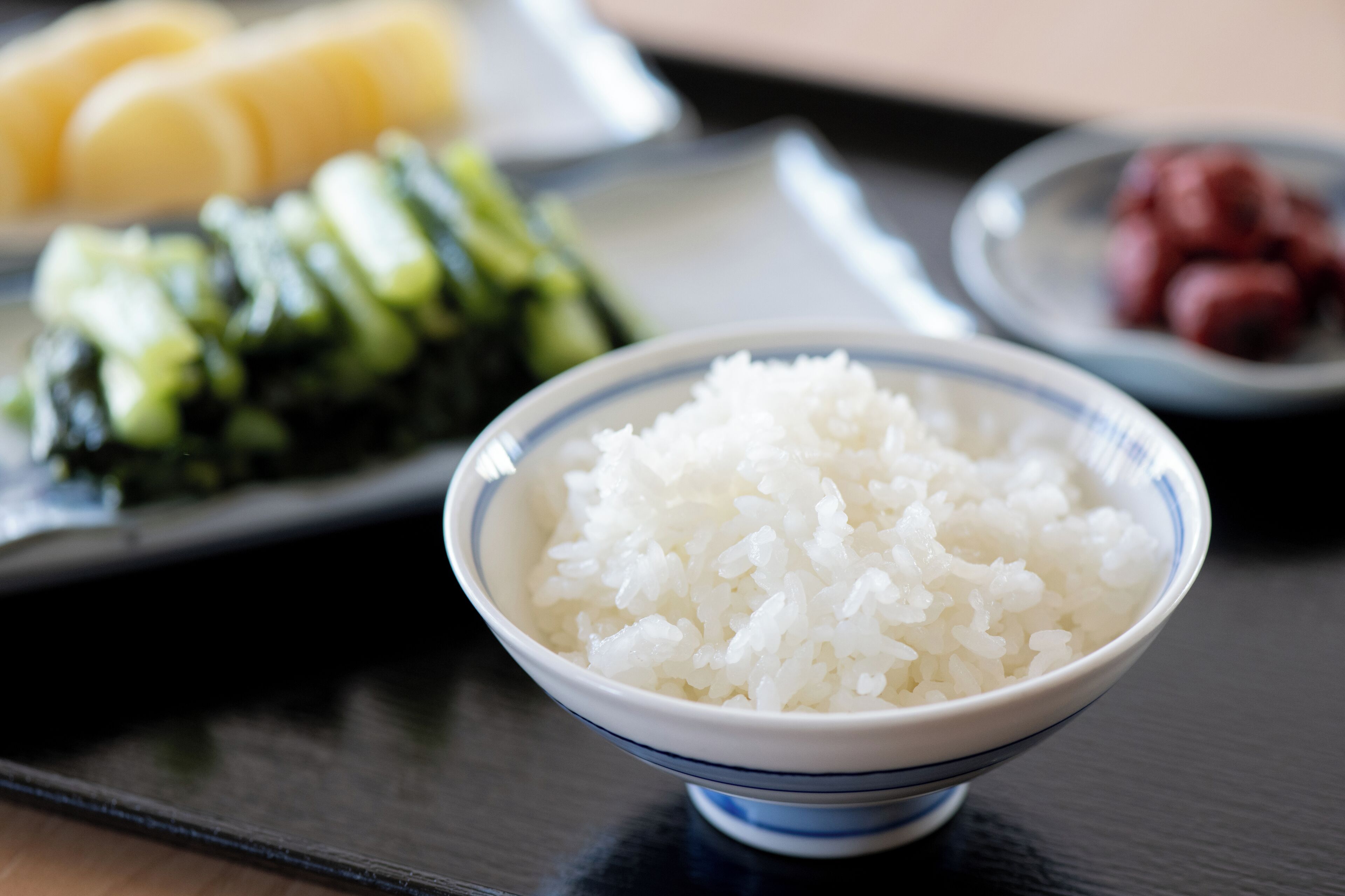 Un bol de riz blanc sur une table, avec une assiette de sushis et une assiette de légumes marinés en arrière-plan.