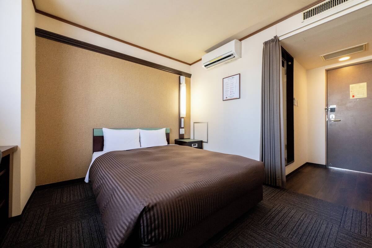 A hotel room with a large bed, a nightstand, a wall-mounted air conditioner, and a door with a handle.