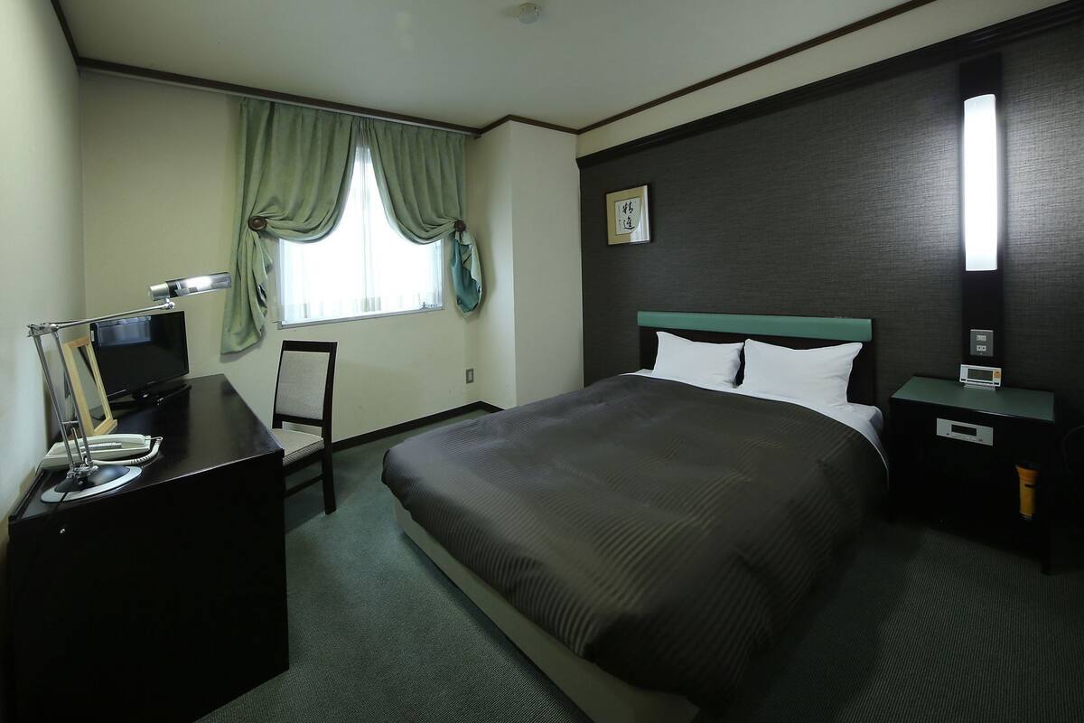 A hotel room with a bed, a desk with a lamp, a chair, a bedside table with a clock, and a window with curtains.