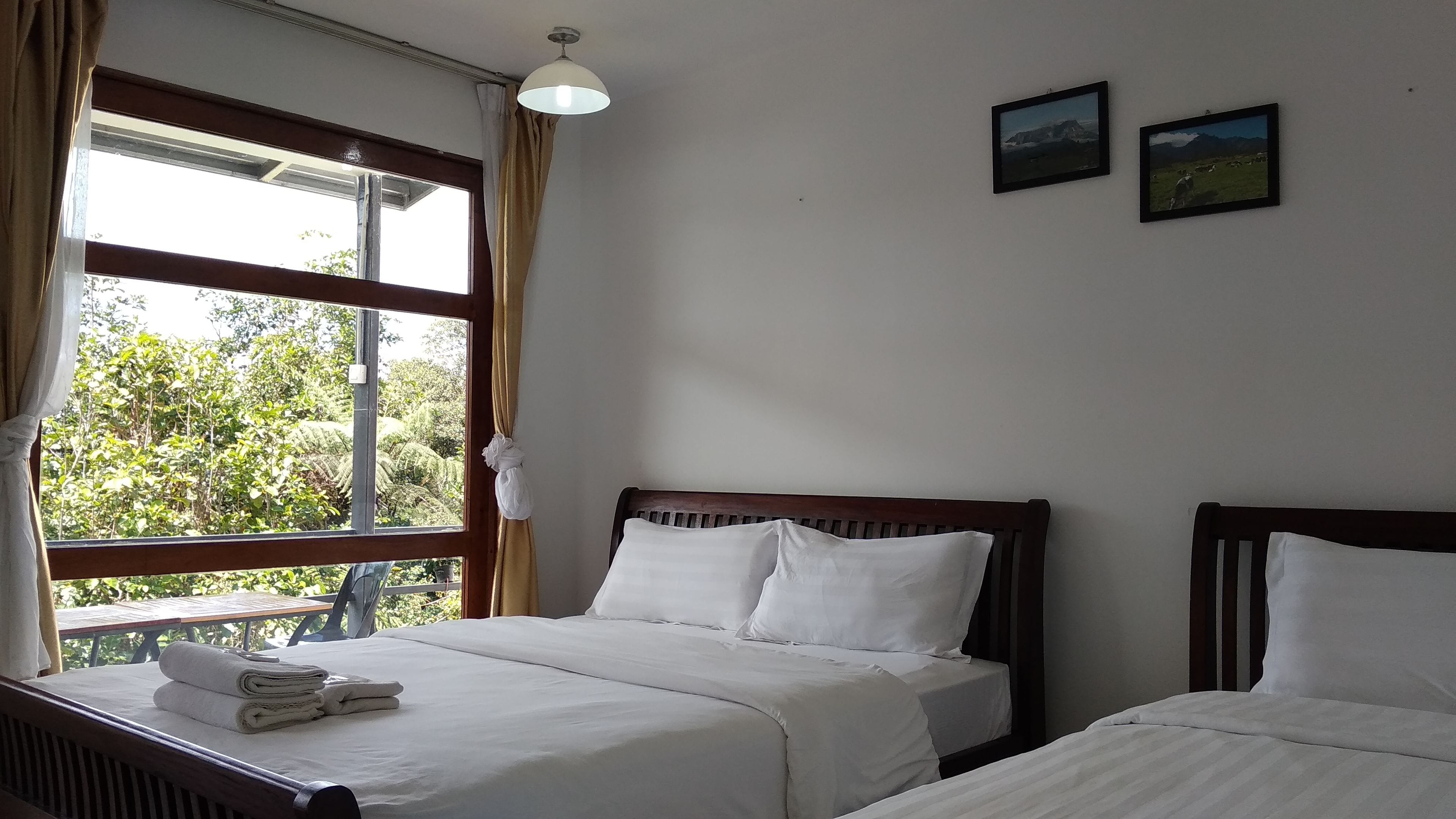 Quarto de hotel com duas camas, uma janela com cortinas, uma luminária no teto e dois quadros na parede.