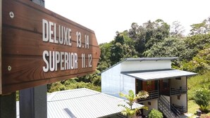 A wooden sign with room names and dates, a modern house with a balcony, and lush greenery in the background.