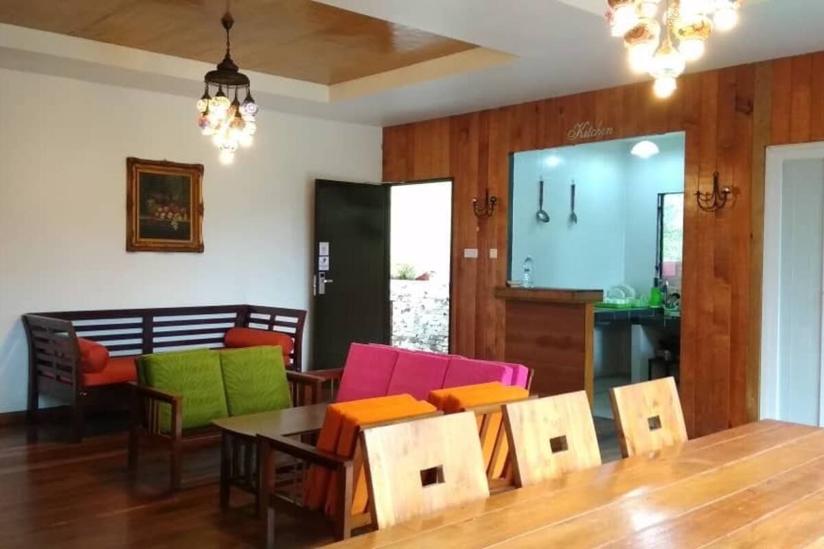 Un coin repas avec une table et des chaises en bois, un canapé agrémenté de coussins colorés et une cuisine visible à travers une porte ouverte.