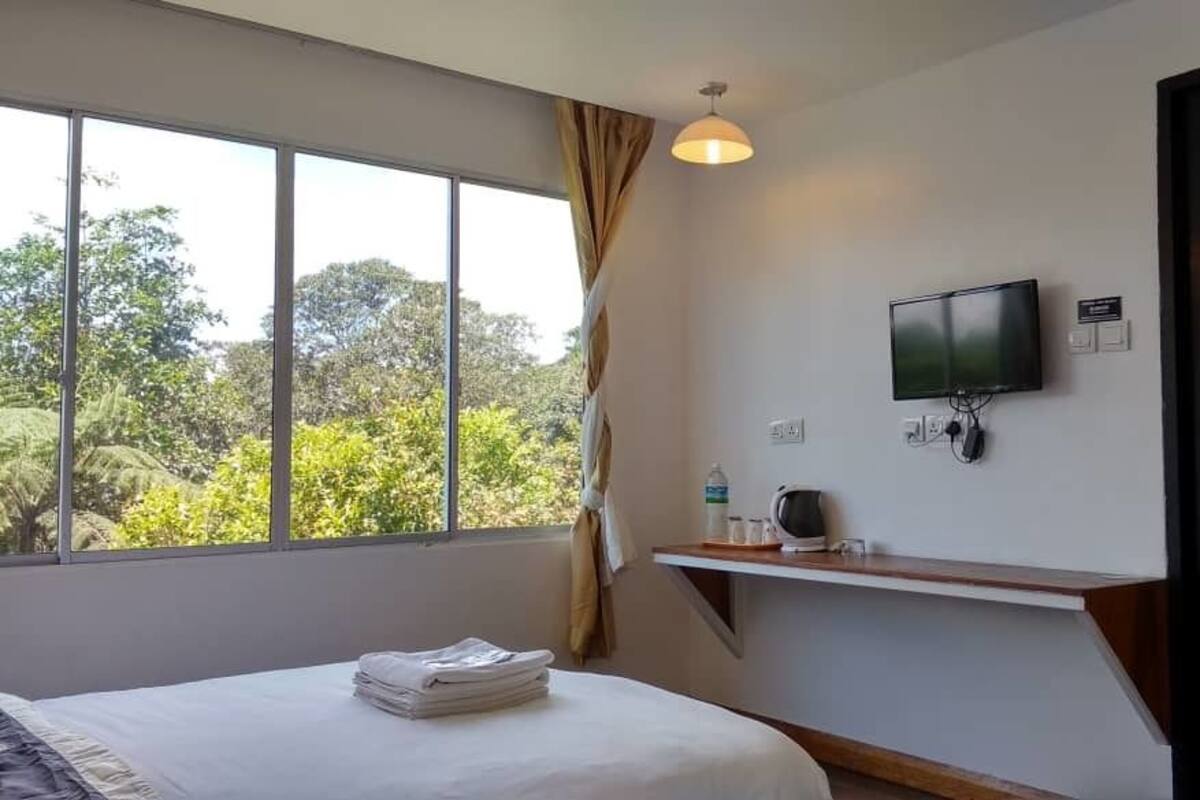 Une chambre d’hôtel avec un lit, un téléviseur fixé au mur, une fenêtre avec des rideaux et une petite étagère avec quelques objets.