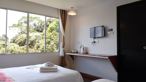 A hotel room with a bed, a TV mounted on the wall, a window with curtains, and a small shelf with a kettle.