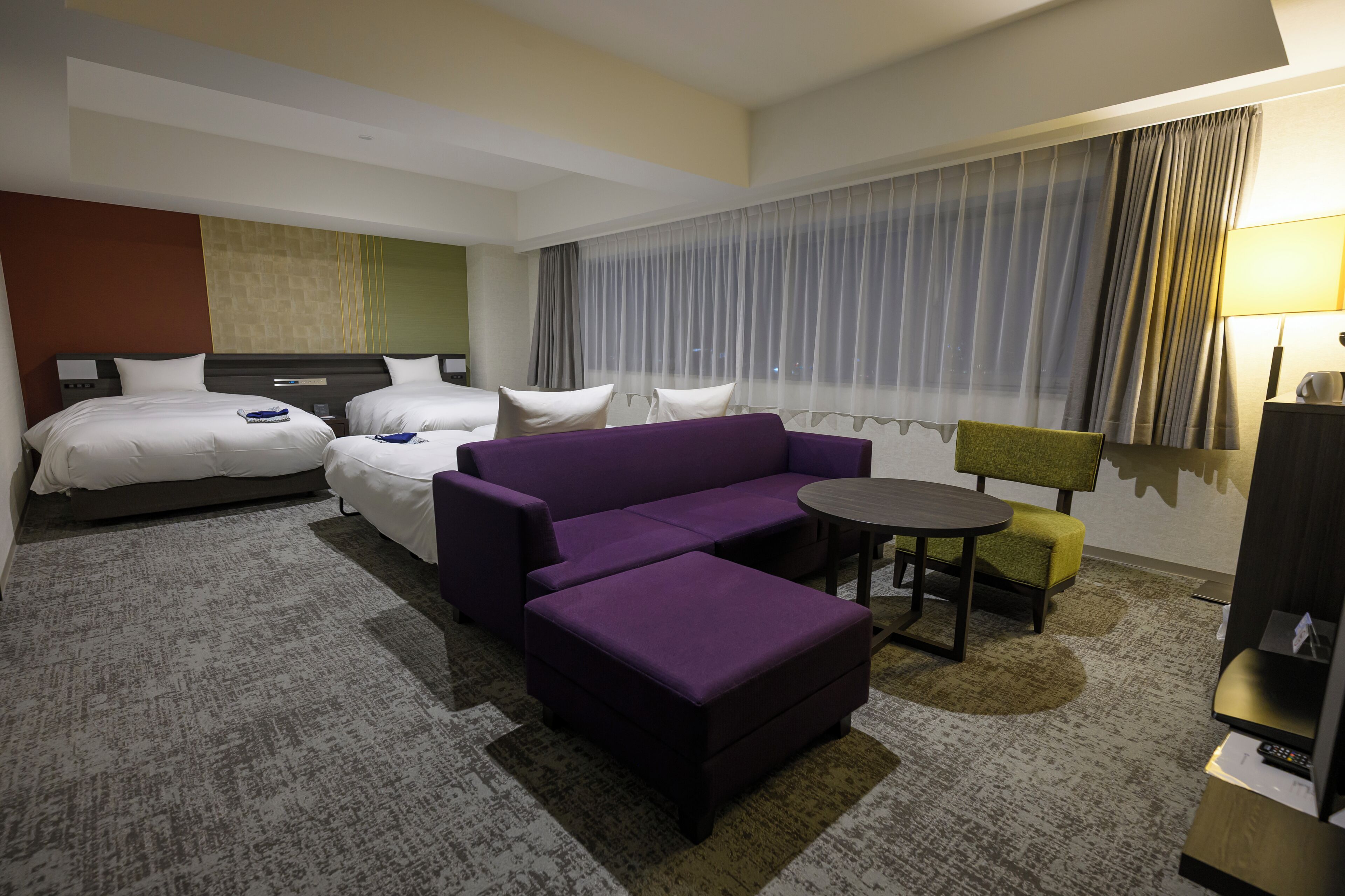 Una habitación de hotel con dos camas, un sofá morado, una mesita y una silla.